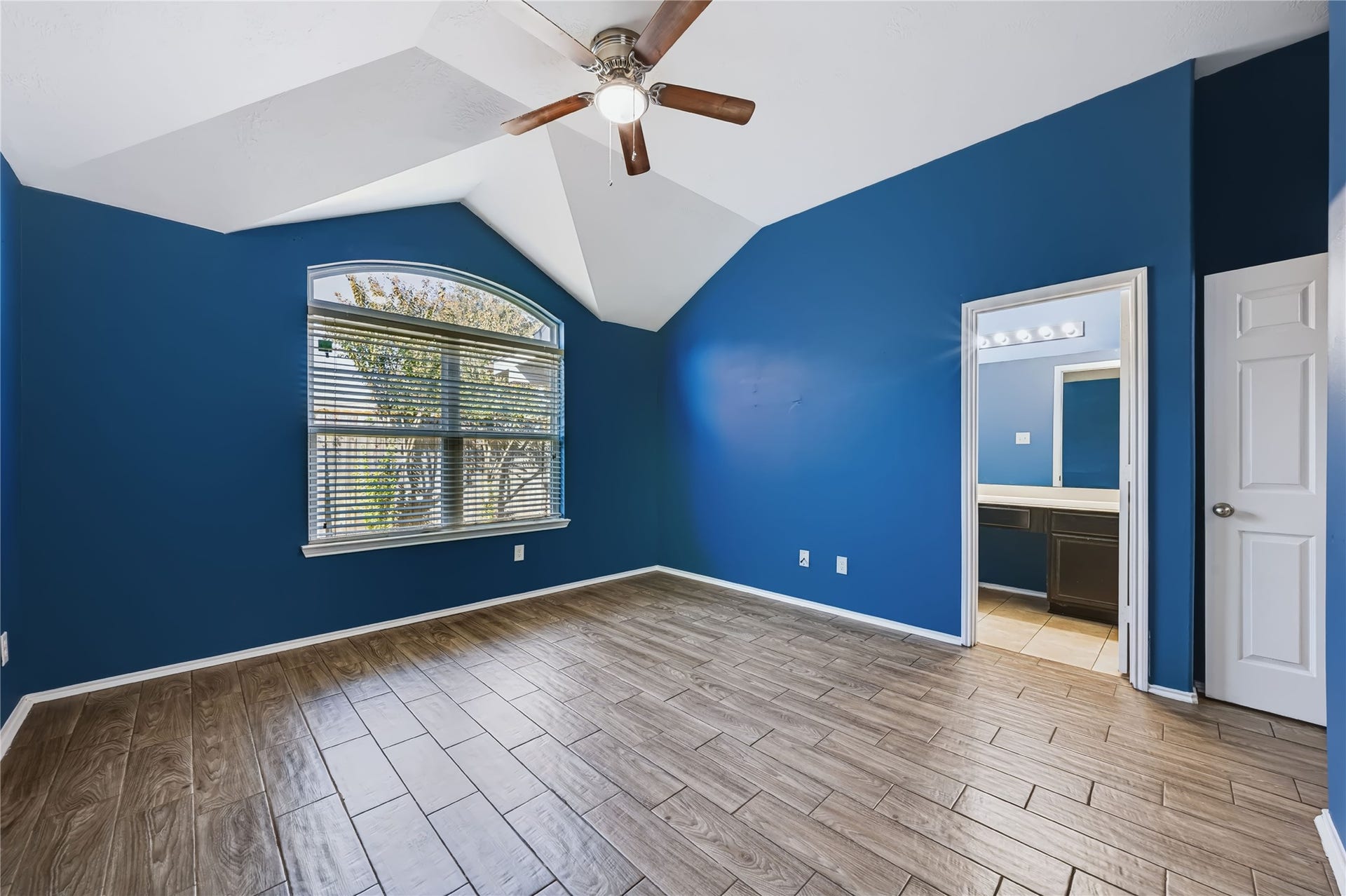 Spacious master bedroom featuring high vaulted ceilings, rich wood-look tile flooring, an upgraded ceiling fan, and large windows bringing in plenty of natural light.