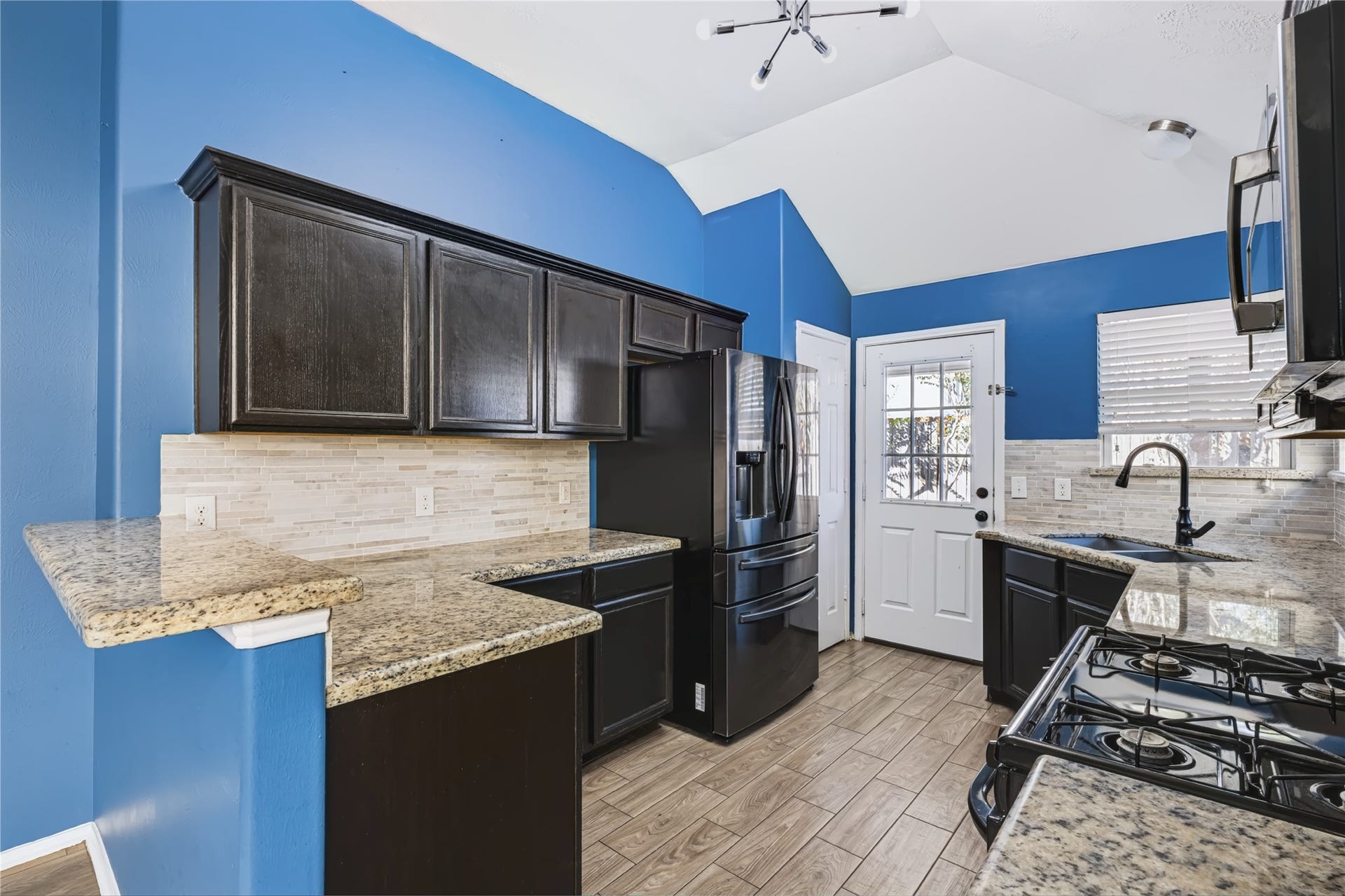 Well-appointed kitchen featuring dark cabinetry, gleaming granite countertops, a gas cooktop, sleek black appliances, and a stylish tiled backsplash with direct access to the backyard.