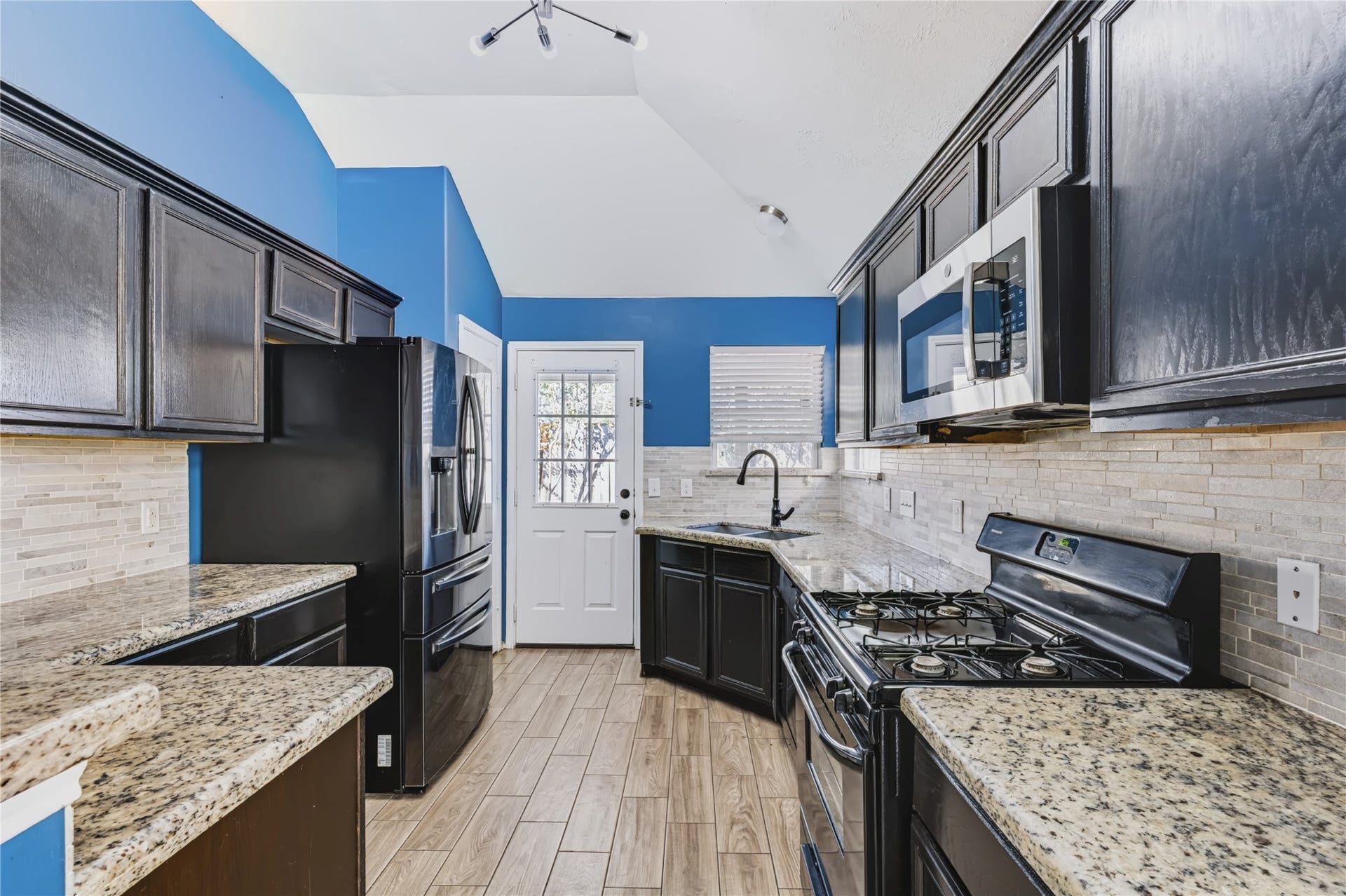 Well-appointed kitchen featuring dark cabinetry, gleaming granite countertops, a gas cooktop, sleek black appliances, and a stylish tiled backsplash with direct access to the backyard.