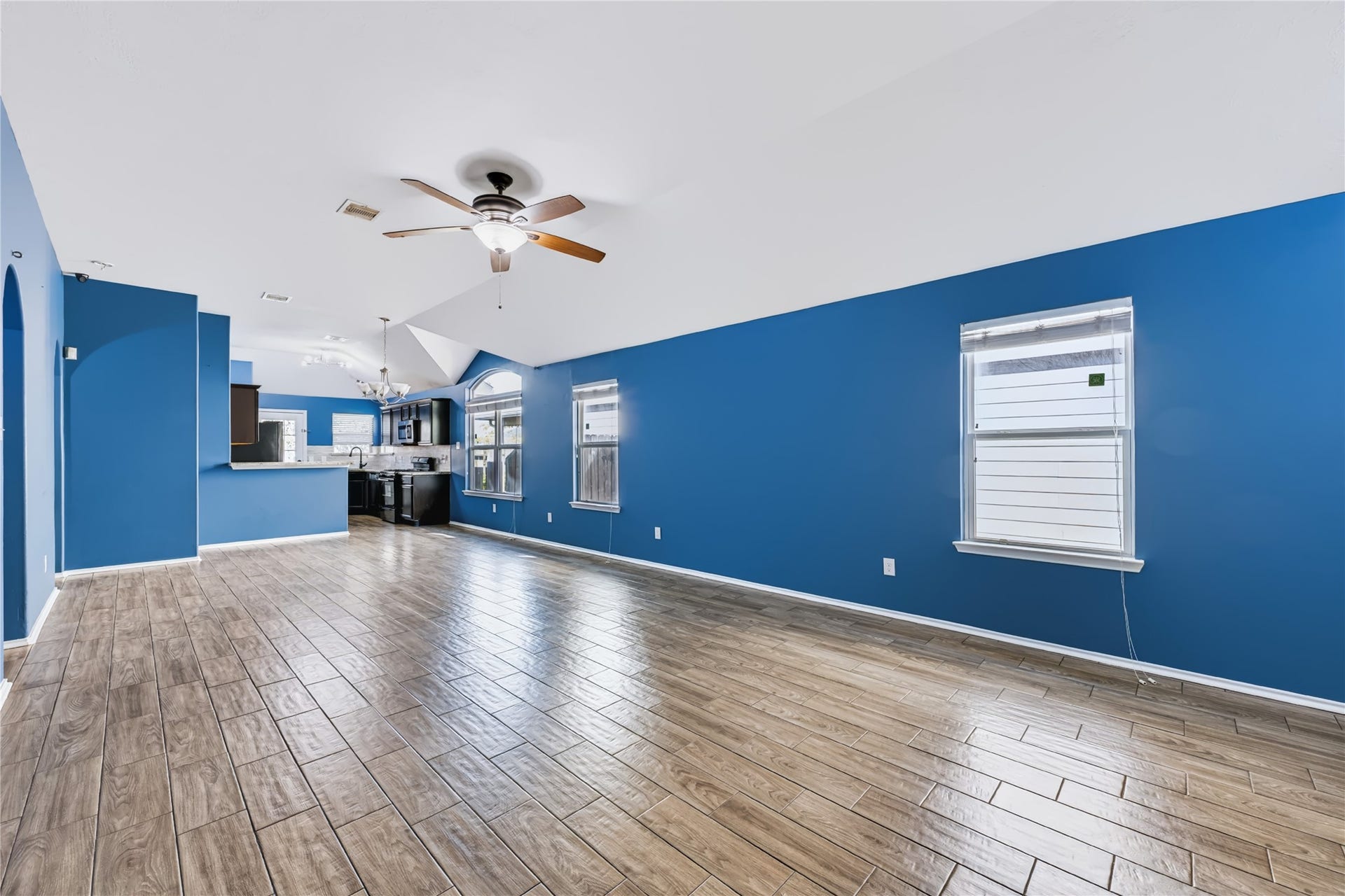 Spacious open-concept living area featuring rich wood-look tile flooring, vaulted ceilings, a modern ceiling fan, and vibrant accent walls that flow seamlessly into the kitchen.