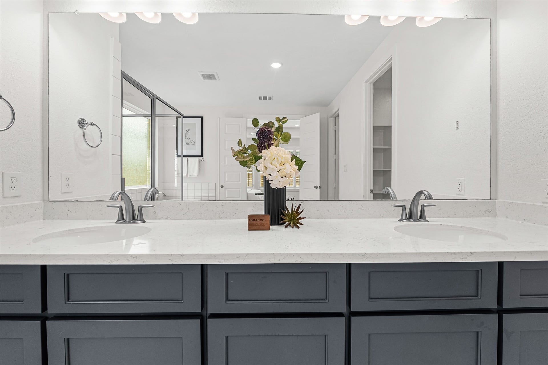 28314 Emerald Bend, Spring, TX 77386 Dark contrast cabinetry and sleek white quartz countertops highlight this primary ensuite dual-sink vanity with brushed nickel fixtures. An expansive wall-to-wall mirror reflects the bright, spa-like atmosphere, including the glass-enclosed shower and freestanding soaking tub across the room.