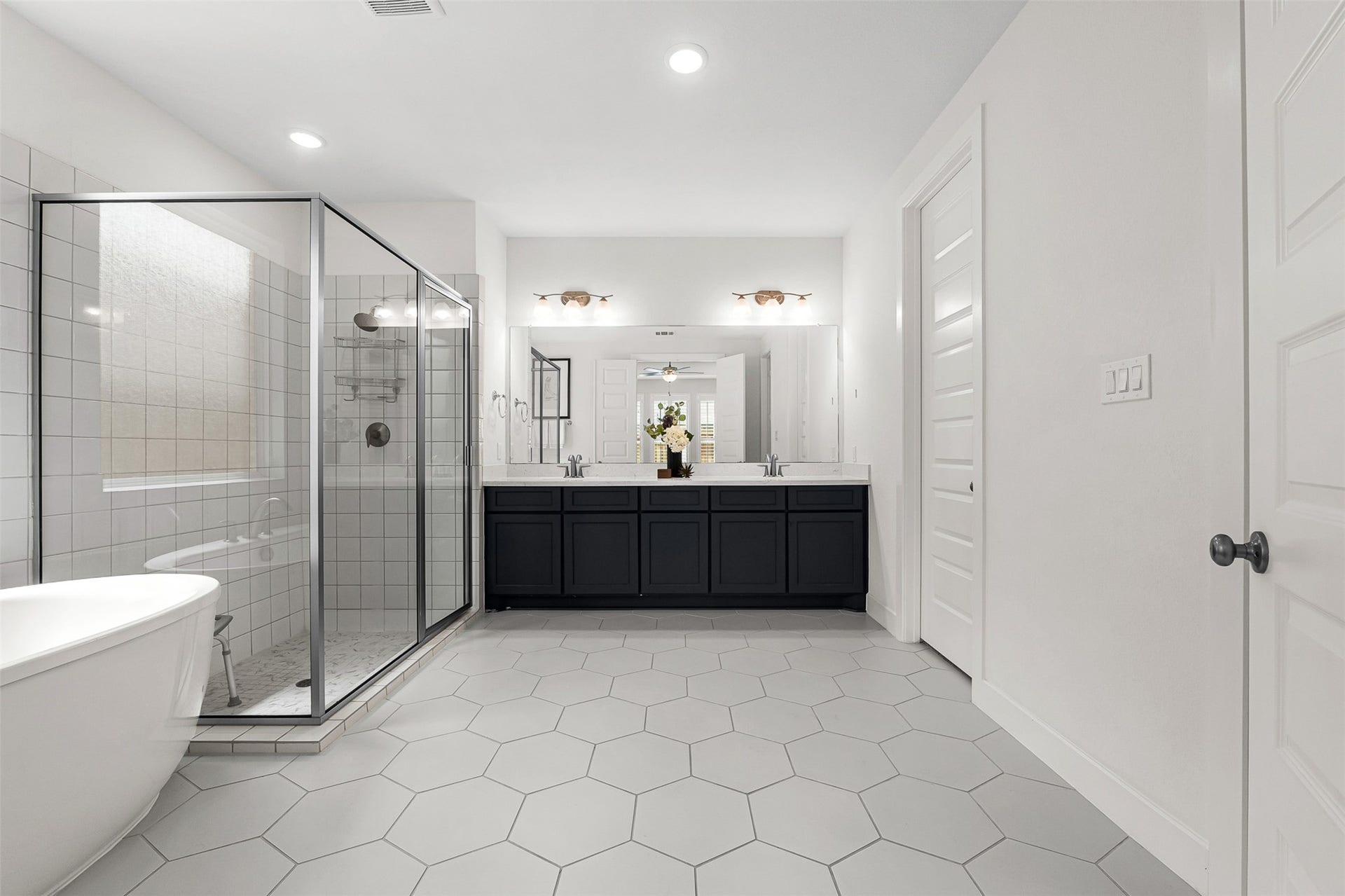 28314 Emerald Bend, Spring, TX 77386 Large-format hexagon tile floors and crisp white walls frame this spa-like primary bath, complete with a freestanding soaking tub and glass-enclosed walk-in shower. A dark contrast vanity with dual sinks, quartz countertops, and classic multi-light sconces provides a striking centerpiece.