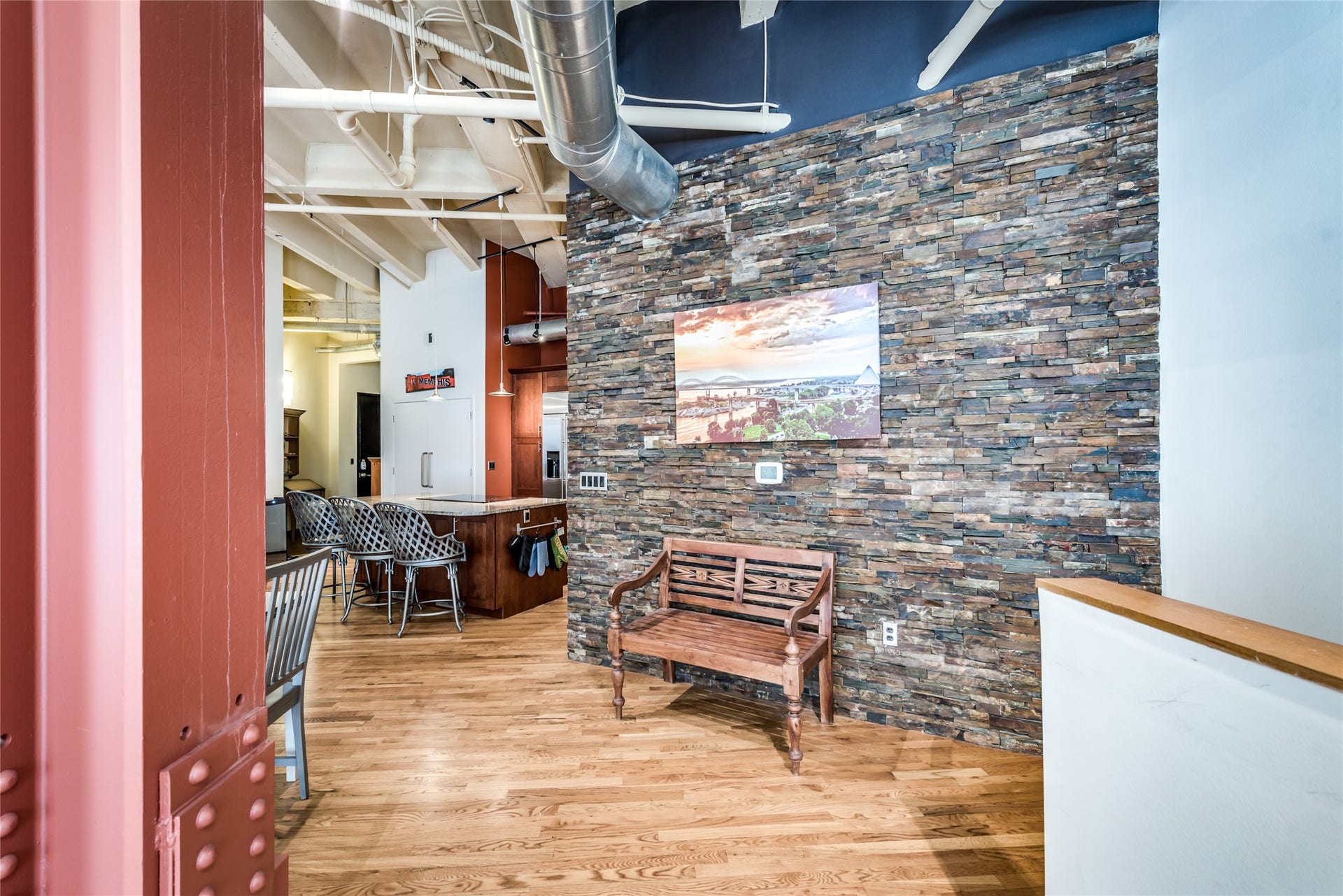 705 Main Street, #215, Houston, TX 77002 This space features a modern industrial design with exposed ductwork and a stone accent wall. The room includes a cozy bench and leads into a kitchen area with a breakfast bar and stylish seating. Warm wood flooring enhances the inviting atmosphere.