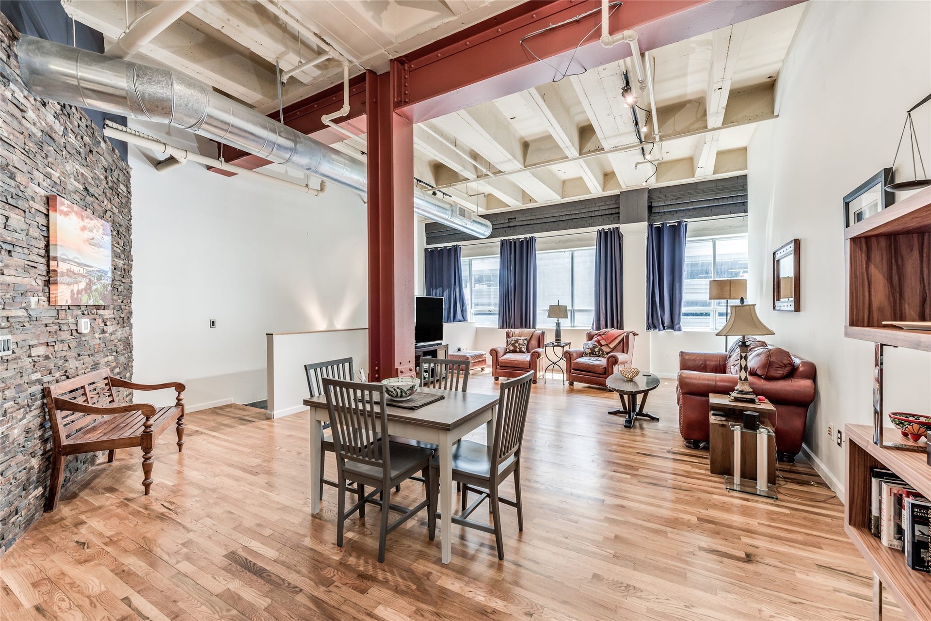 705 Main Street, #215, Houston, TX 77002 This photo showcases a spacious, open-concept loft with high ceilings, exposed ductwork, and a mix of industrial and cozy elements. The room features polished wood floors, a brick accent wall, and large windows with ample natural light. The living area includes comfortable seating and a dining space, perfect for modern urban living.