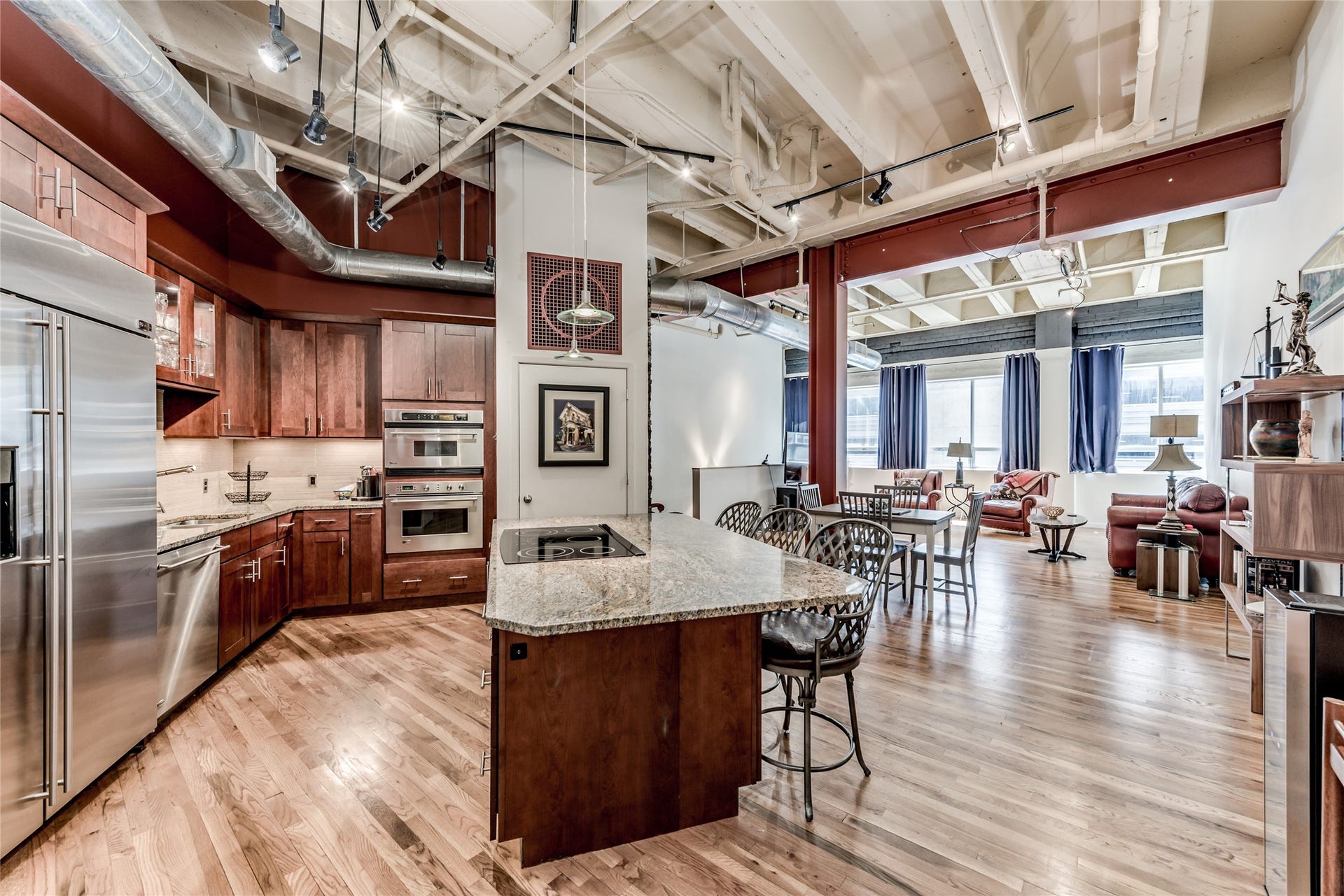 705 Main Street, #215, Houston, TX 77002 This photo showcases a spacious, modern open-concept living area with a sleek kitchen featuring stainless steel appliances, a central island with cook top and breakfast bar seating, and rustic wood cabinetry. The space extends into a light-filled living and dining area, highlighting high ceilings with exposed beams and ductwork, creating an industrial yet inviting atmosphere.