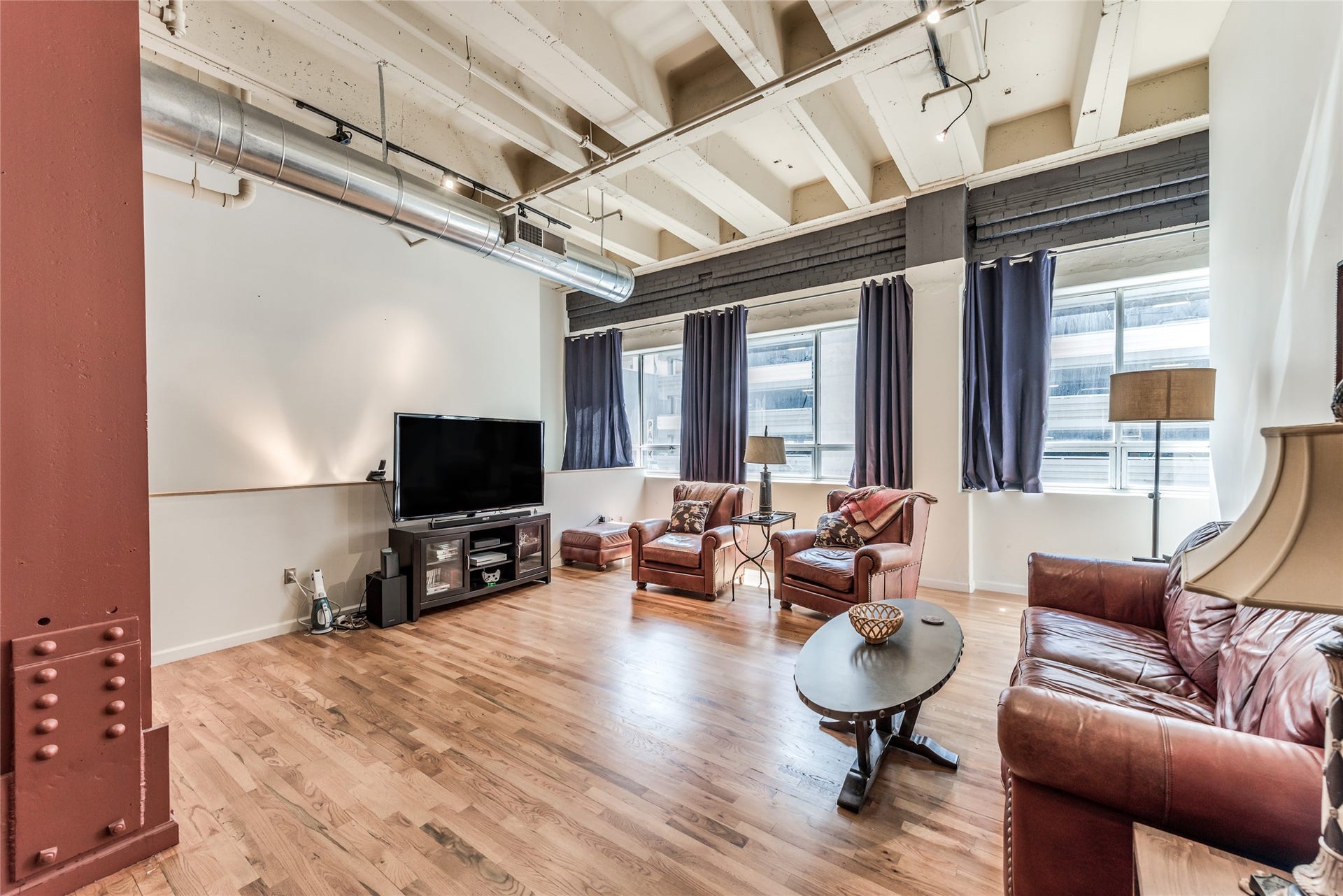 705 Main Street, #215, Houston, TX 77002 This photo showcases a spacious, industrial-style living room with high ceilings, exposed beams, and ductwork. It features large windows, hardwood floors, leather furniture, and a cozy seating area with a TV. The space is bright and modern, ideal for comfortable living.