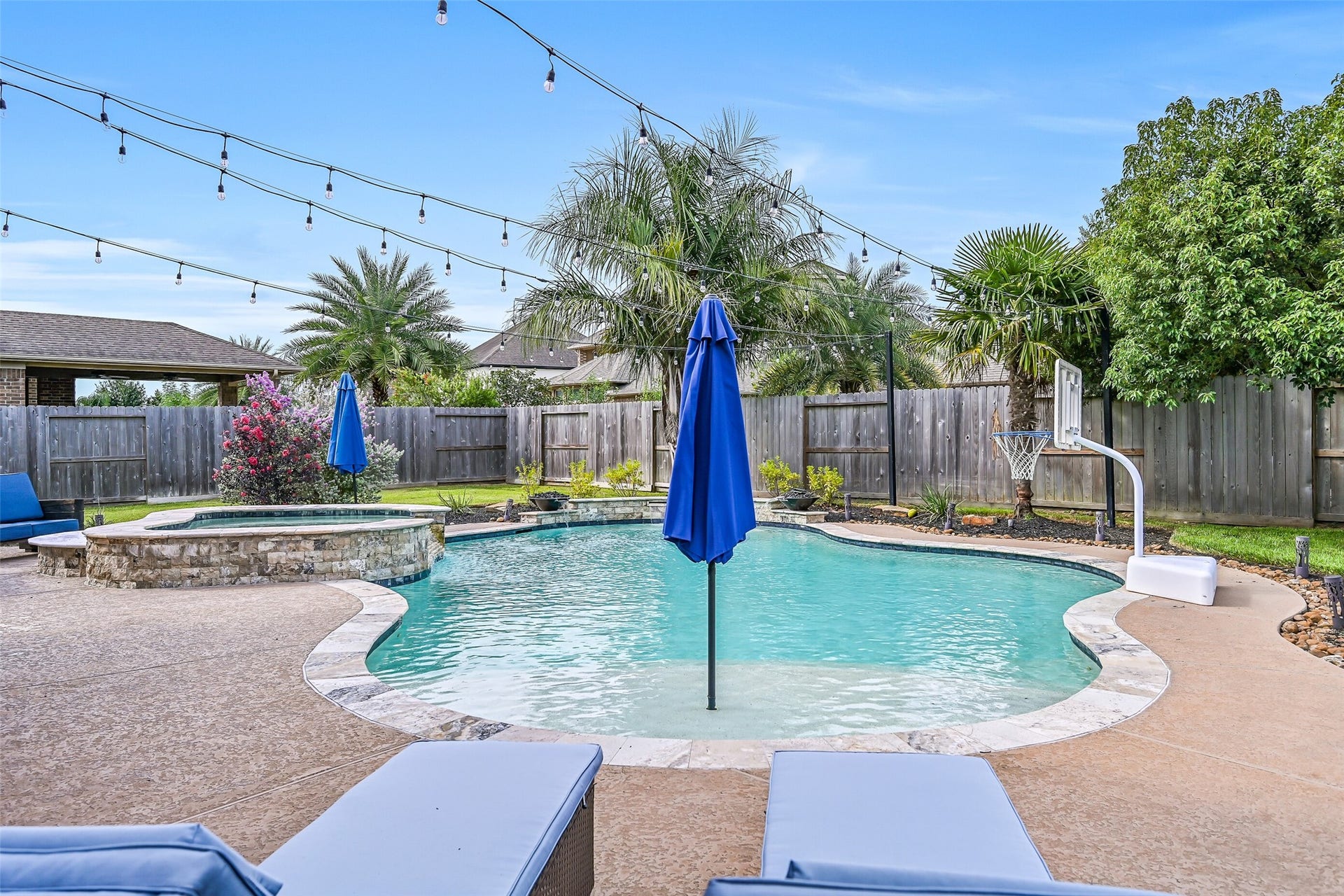 A closer view showcases the freeform heated pool, oversized raised spa with spillover, travertine coping and surrounding landscape beds, all accented by overhead string lights and landscape lighting.