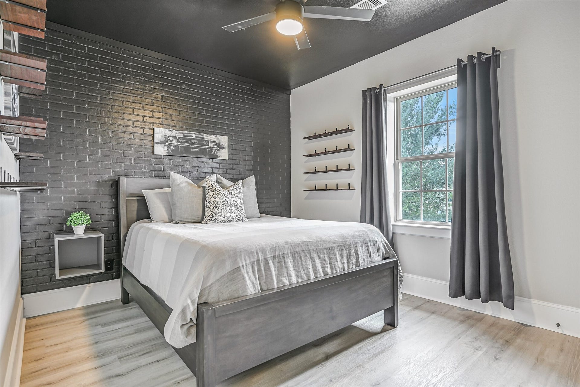 This upstairs secondary bedroom features LVP flooring, a dramatic black-painted brick accent wall, black ceiling, lighted ceiling fan and industrial-style shelving. A walk-in closet and Jack & Jill bath connect it to the adjoining bedroom.