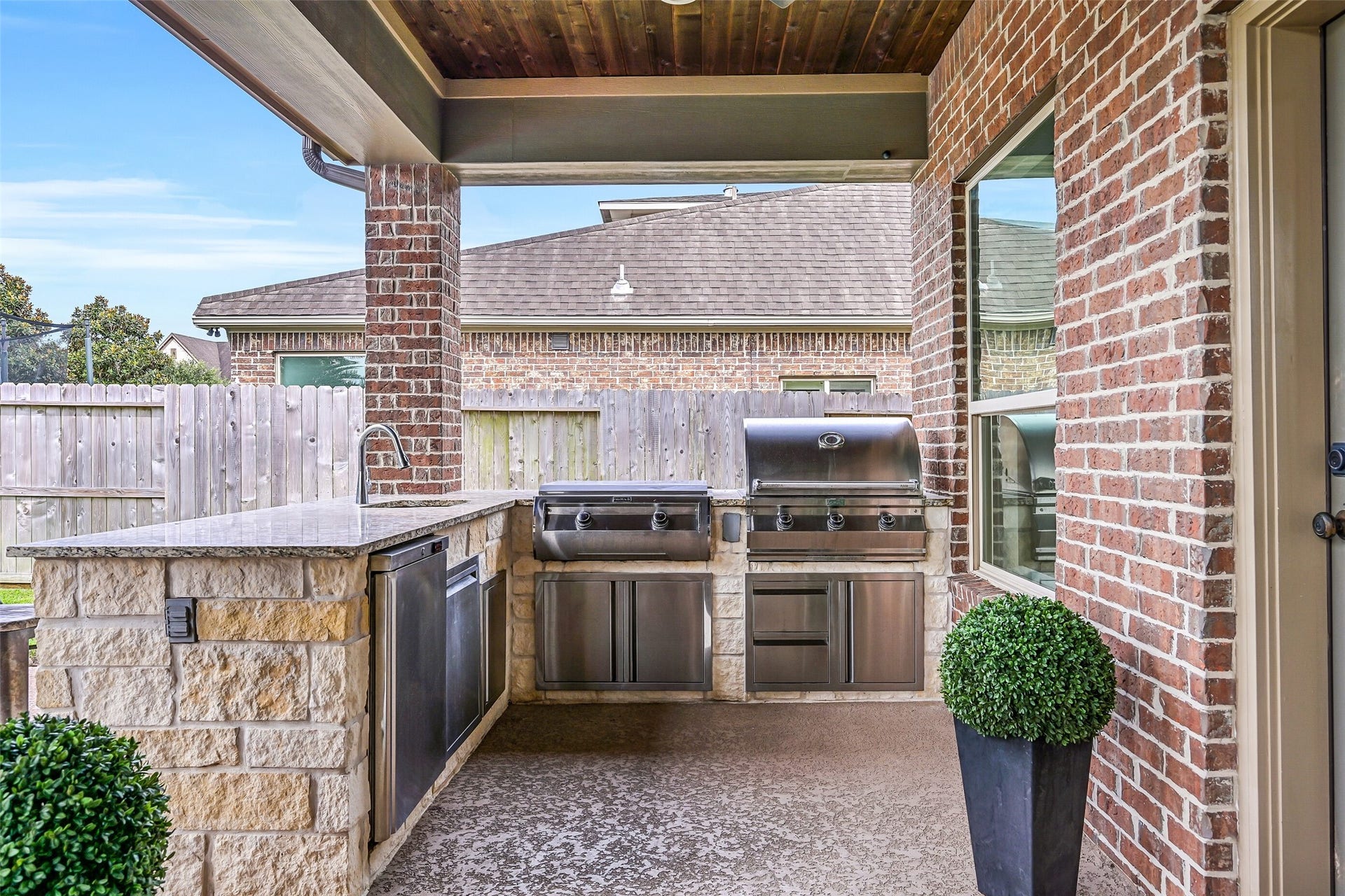 A fully equipped outdoor kitchen features granite counters, stone surround, beverage refrigerator, bar sink, stainless storage, Fire Magic flat-top grill and Aurora gas grillall tucked beneath the covered patio.