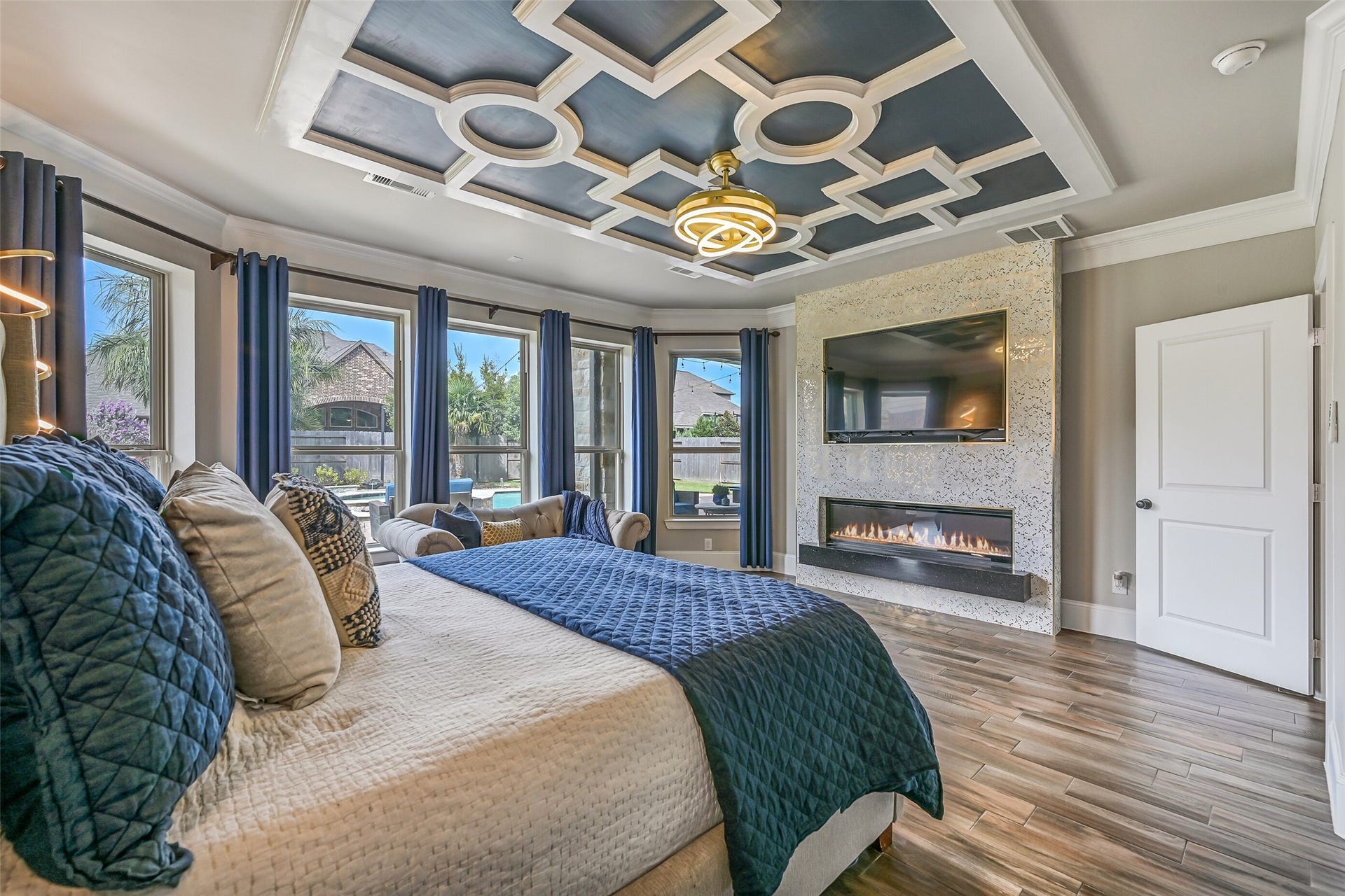 The secluded first-floor primary retreat makes a statement with an elaborate custom ceiling treatment, modern lighting, wood-look tile floors & a sleek linear electric fireplace with full-height surround, while a wall of windows overlooks the backyard pool.