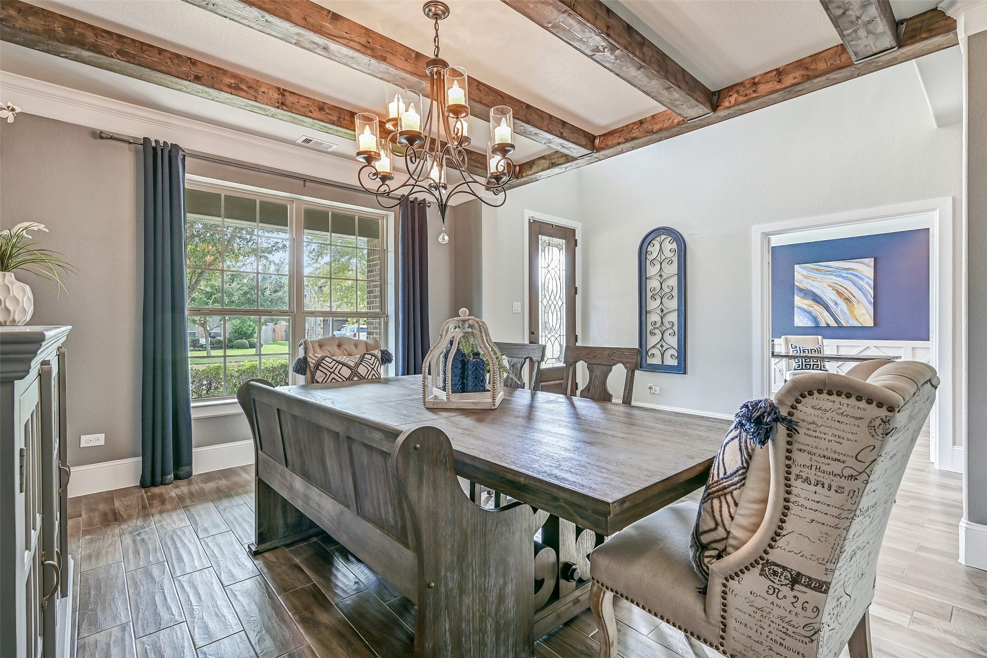 The striking dining room showcases four substantial wood ceiling beams, crown molding, wood-look tile flooring, a stylishly updated chandelier and custom wall treatments in a sophisticated navy-and-neutral palette.