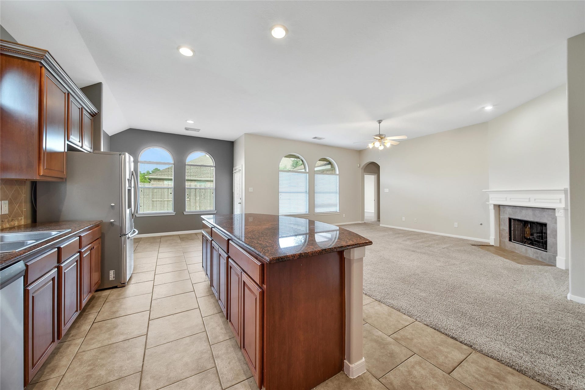 25604 Southwood Oaks Court, Porter, TX 77365 This is a spacious, open-plan living area with a modern kitchen featuring stainless steel appliances, wooden cabinets, and a central island. The room transitions into a cozy carpeted living space with ample natural light, a fireplace, and a view of the outdoors through large windows.