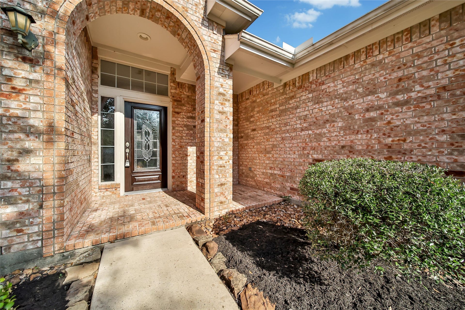25604 Southwood Oaks Court, Porter, TX 77365 This is an image of a home's welcoming entrance, featuring a brick archway leading to a wood front door with decorative glass and a sidelight. The home has a clean, well-maintained appearance with a neat landscaping touch on the side.
