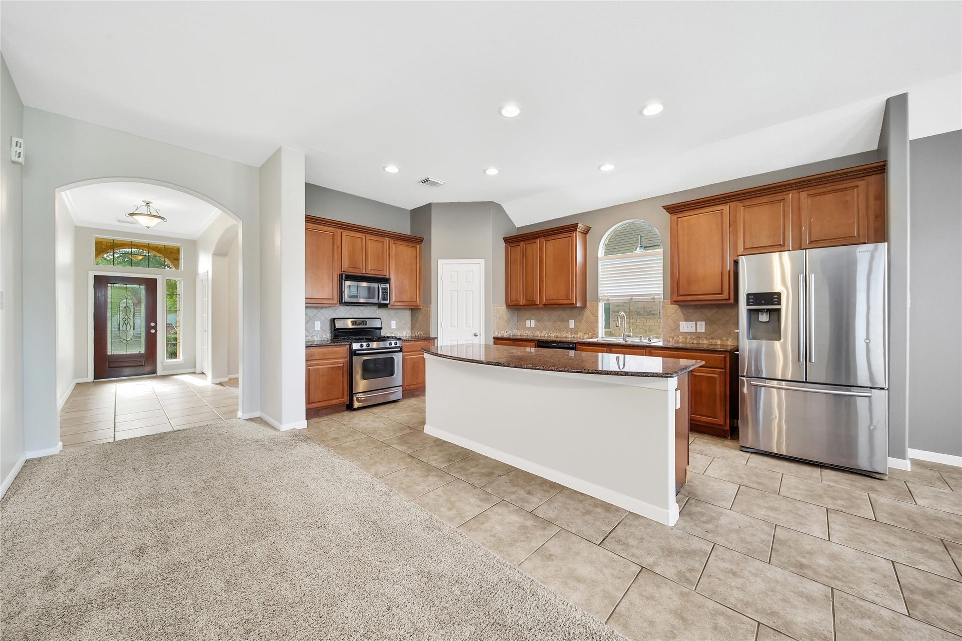 25604 Southwood Oaks Court, Porter, TX 77365 You're looking at a spacious kitchen with modern stainless steel appliances, ample wooden cabinetry, and granite countertops. The kitchen opens up to a welcoming entryway with a view of the front door through an arched passageway. The room features a blend of tile flooring in the kitchen area and plush carpeting in the adjoining space.
