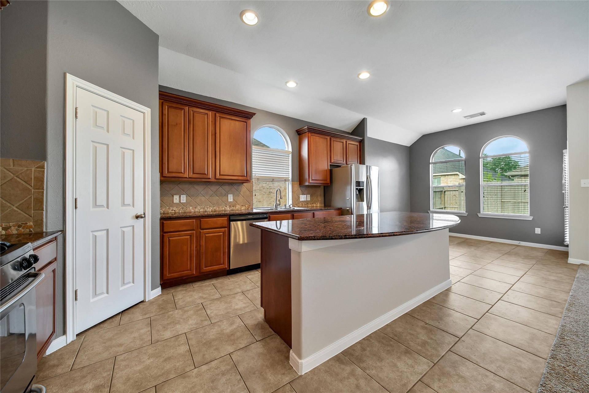 25604 Southwood Oaks Court, Porter, TX 77365 This is a spacious kitchen featuring rich wooden cabinetry, modern stainless-steel appliances, tiled flooring, and a central island with countertop space. Natural light streams in through arched windows, complementing the neutral color palette and recessed lighting.