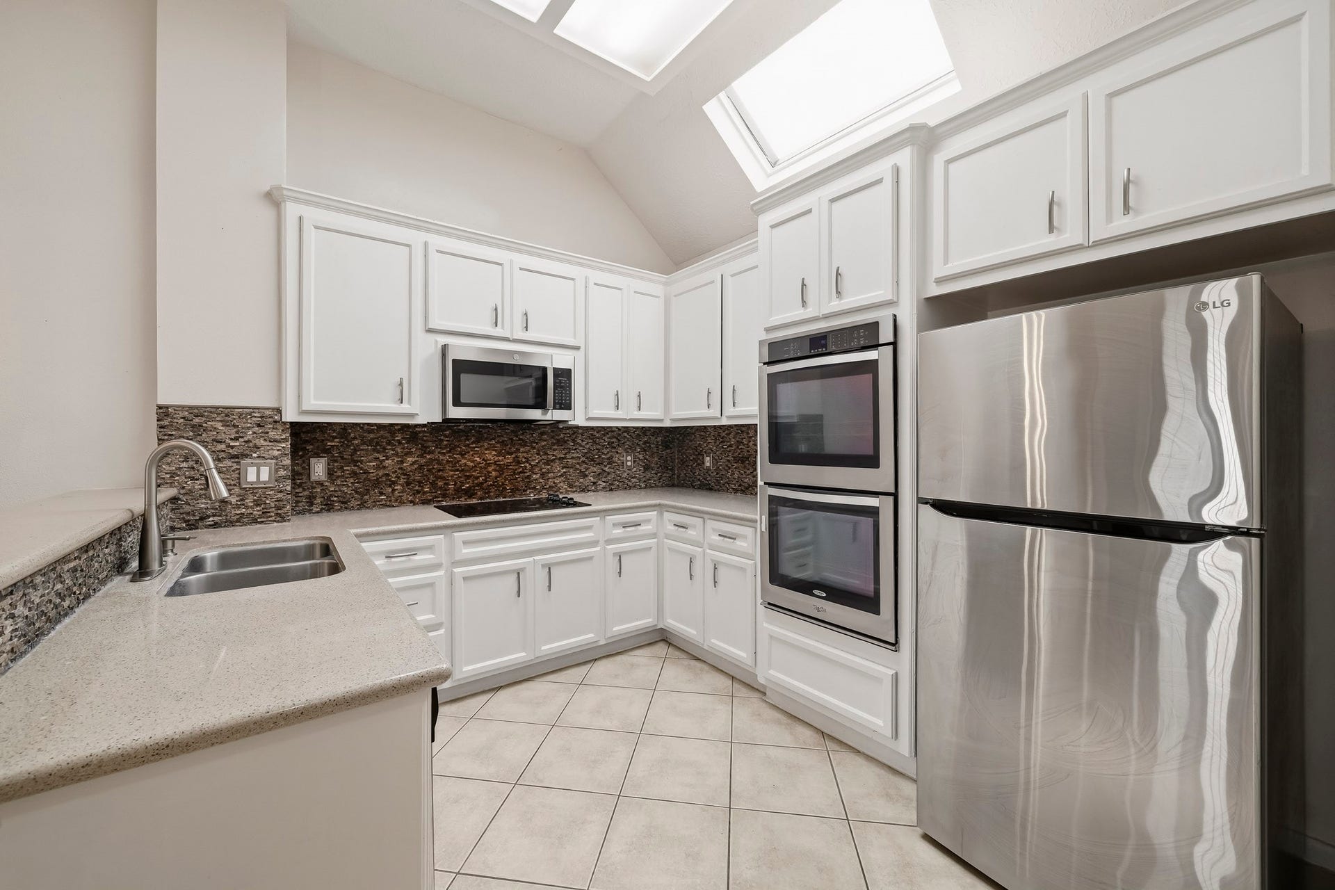 2611 Oxhill Court, Pearland, TX 77584 This bright kitchen features crisp white cabinetry, generous counter space, and a striking mosaic tile backsplash. Stainless steel appliances include a refrigerator, built-in double ovens, microwave, and gas cooktop. Multiple skylights bring abundant natural light into the space, while the wraparound layout and breakfast bar provide plenty of room for meal preparation, storage, and everyday entertaining.
