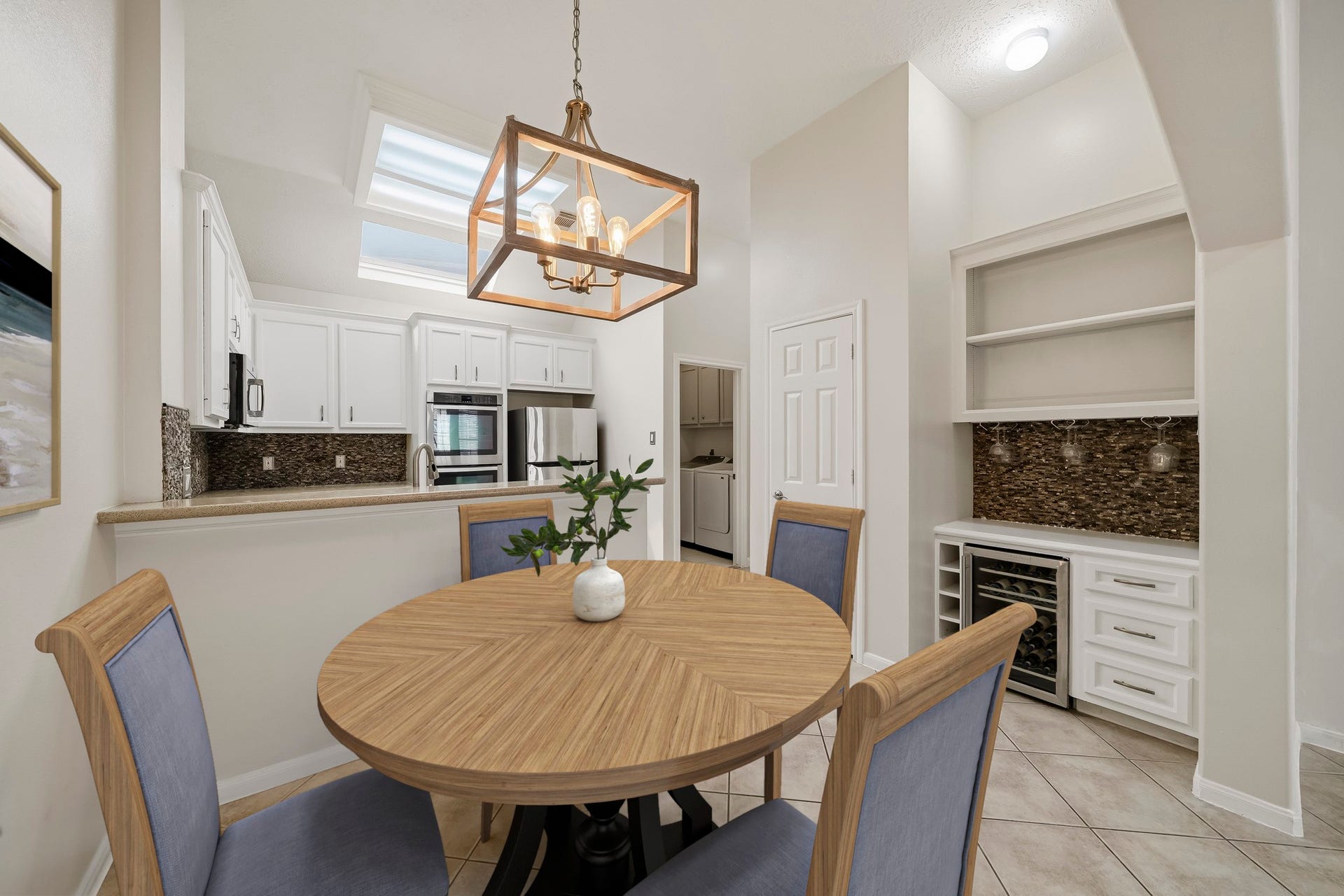 2611 Oxhill Court, Pearland, TX 77584 Virtually Staged: This bright breakfast area offers an open connection to the kitchen, highlighted by a large breakfast bar and a striking pendant chandelier. The space features tile flooring, and a built-in beverage station with cabinetry, shelving, wine storage, and a beverage refrigerator. Skylights and neutral finishes enhance the light, open feel, while the nearby laundry room adds everyday convenience.