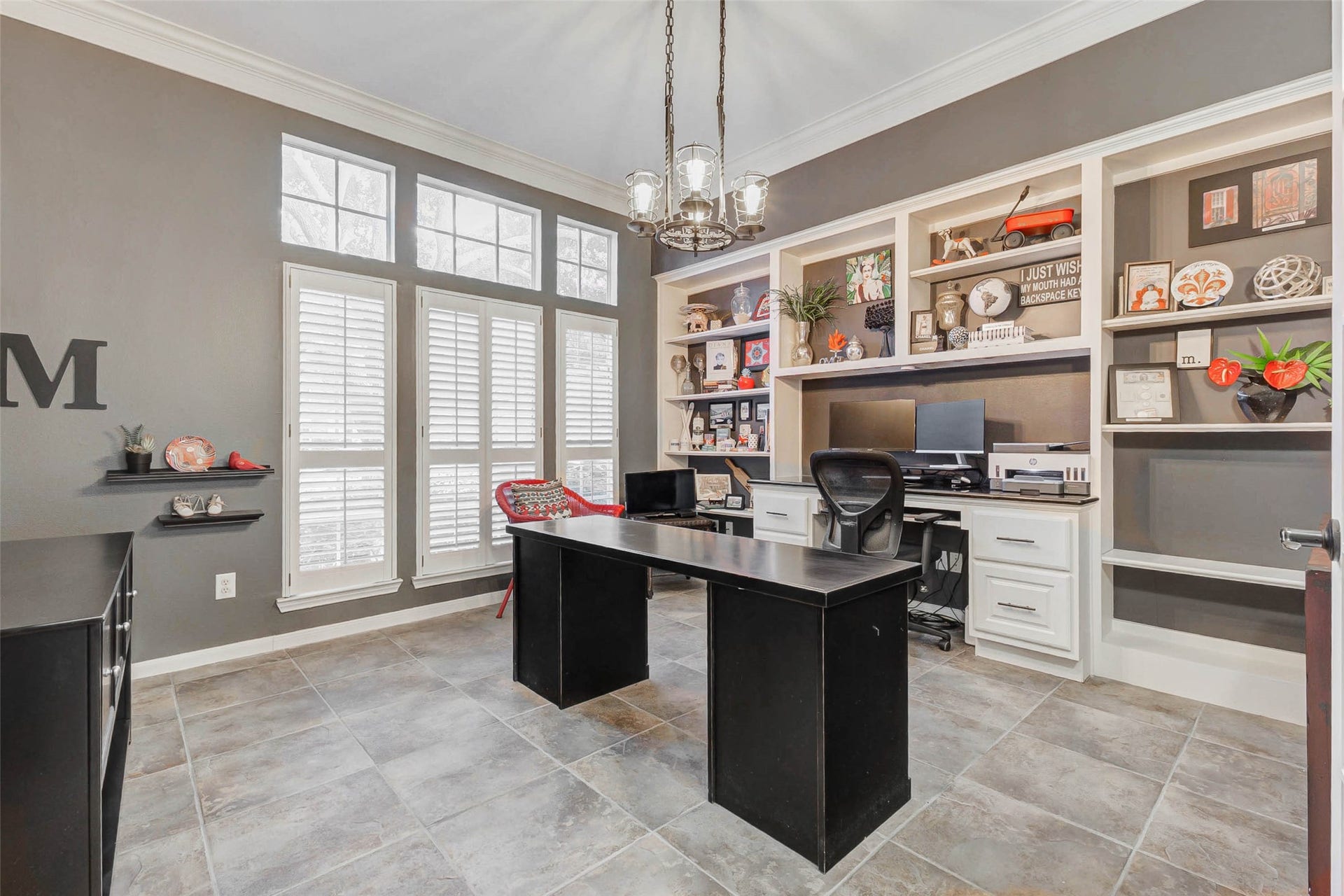 This photo showcases a bright home office with large windows featuring plantation shutters, built-in shelves, desk with granite top, and a modern chandelier. The space is accented with neutral tones and offers ample room for productivity.