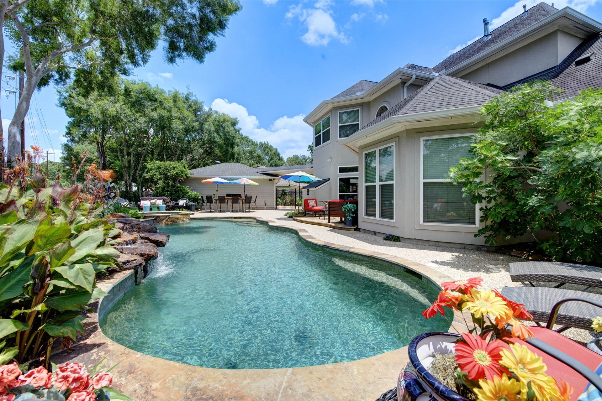 This photo features a beautiful two-story home with a spacious backyard showcasing a custom swimming pool surrounded by lush landscaping. There are seating areas with umbrellas, perfect for relaxing and entertaining, and the exterior is light-colored with large windows, providing plenty of natural light.