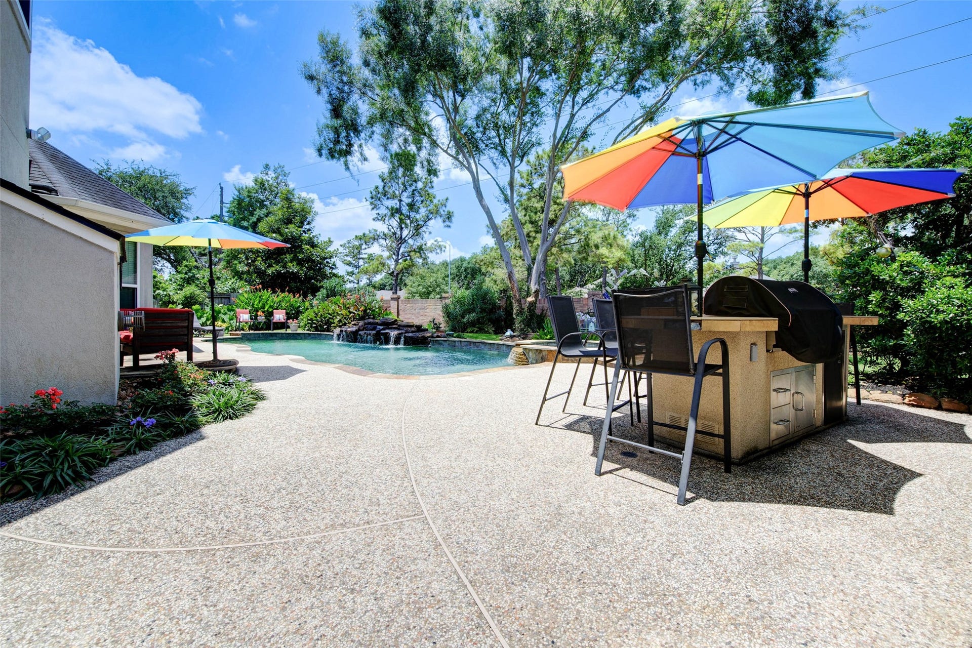 This inviting backyard features a sparkling pool with a waterfall, surrounded by lush greenery. There's a spacious patio with outdoor kitchen with bar seating and 2 umbrellas holes, 2 drawers, runs off of propane tank, plug underneath kitchen for refrigerator and one electrical outlet on the kitchen itself. Additional burner. Bull grill with 6 burners. Mini refrigerator in the outdoor kitchen comes with the house too. Perfect for outdoor entertaining and relaxation.