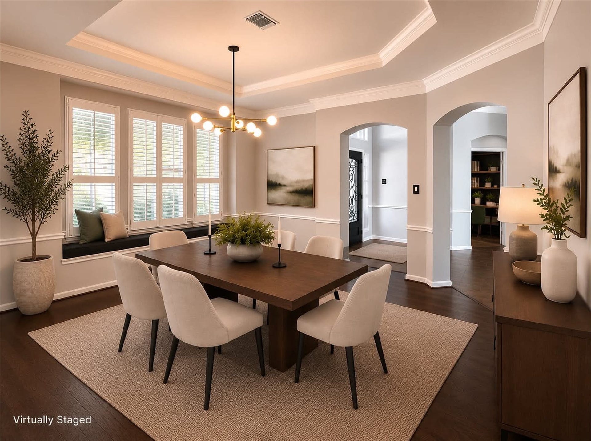 *virtually staged* photo showcases a stylish dining room with elegant light gray walls and white trim. It features a modern chandelier, large windows with shutters. The space is accented with dark wood flooring, offering a blend of sophistication and comfort. Custom crown molding that also continues throughout the house.