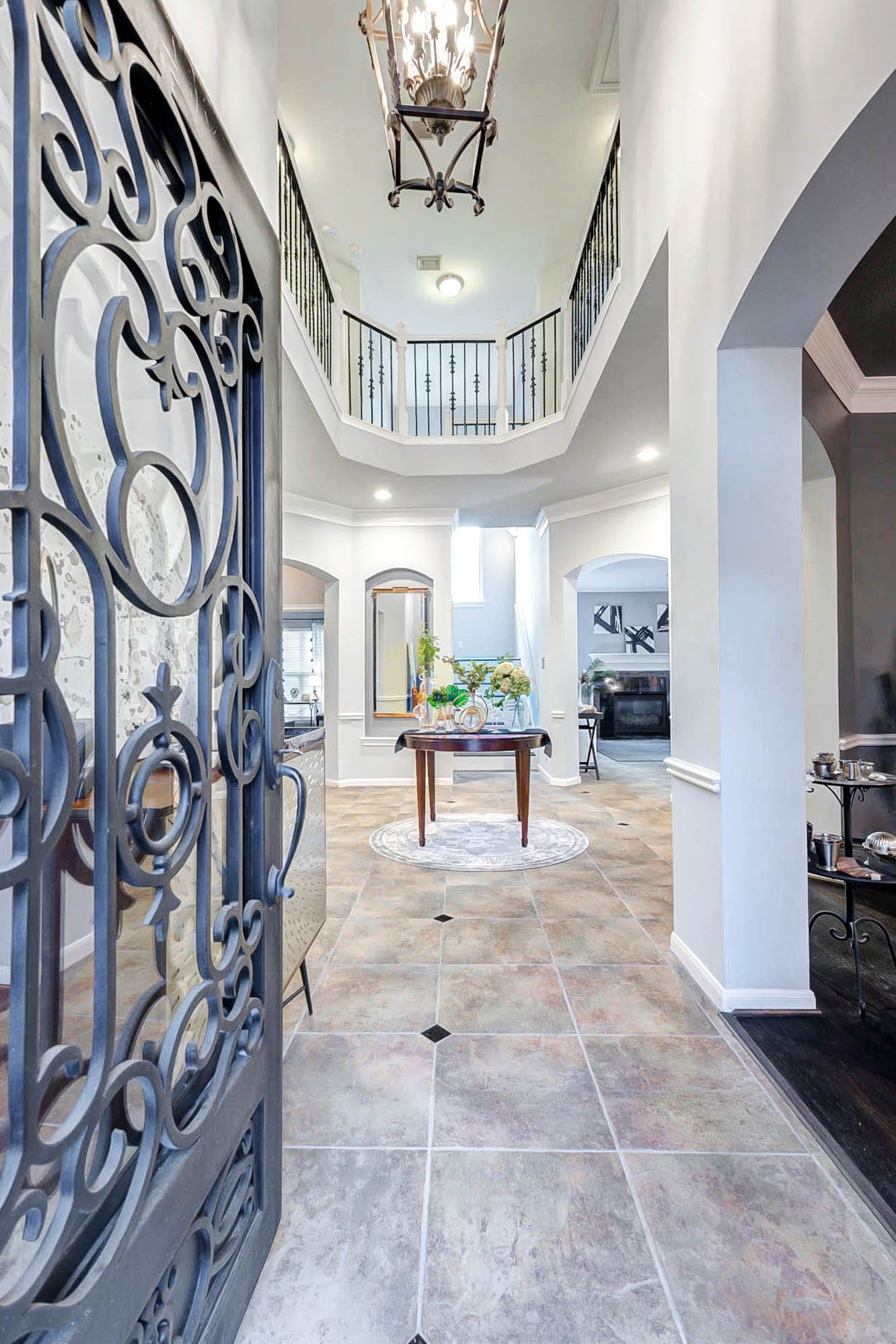 This photo showcases a grand entryway with a decorative wrought iron door, high ceilings, and elegant lighting. The space features tile flooring and an open view to the second floor, creating a spacious, inviting atmosphere.