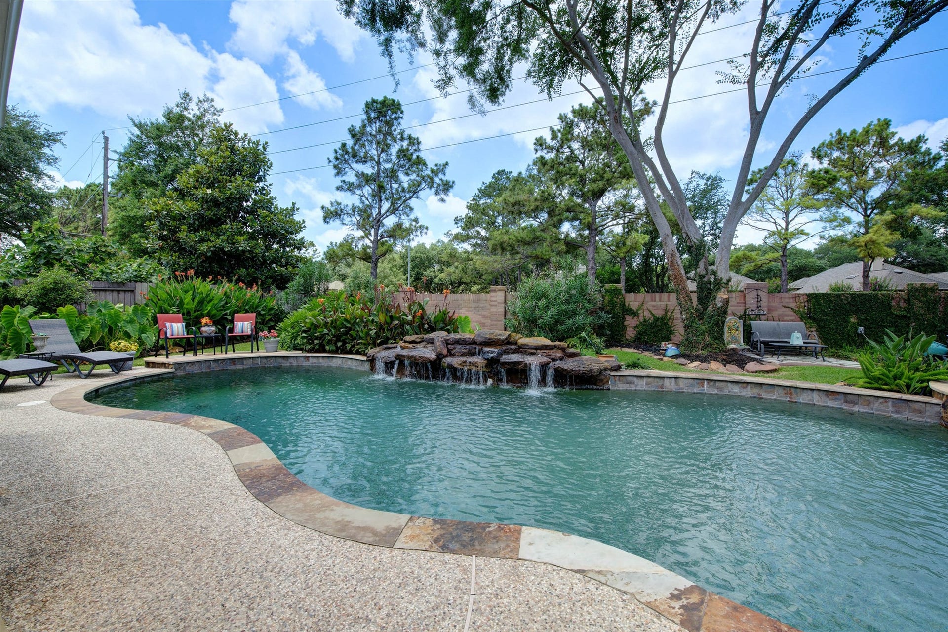 This photo showcases a serene backyard oasis featuring a curvy swimming pool with a rock waterfall feature. It's surrounded by lush greenery and mature trees, offering a private and tranquil retreat.