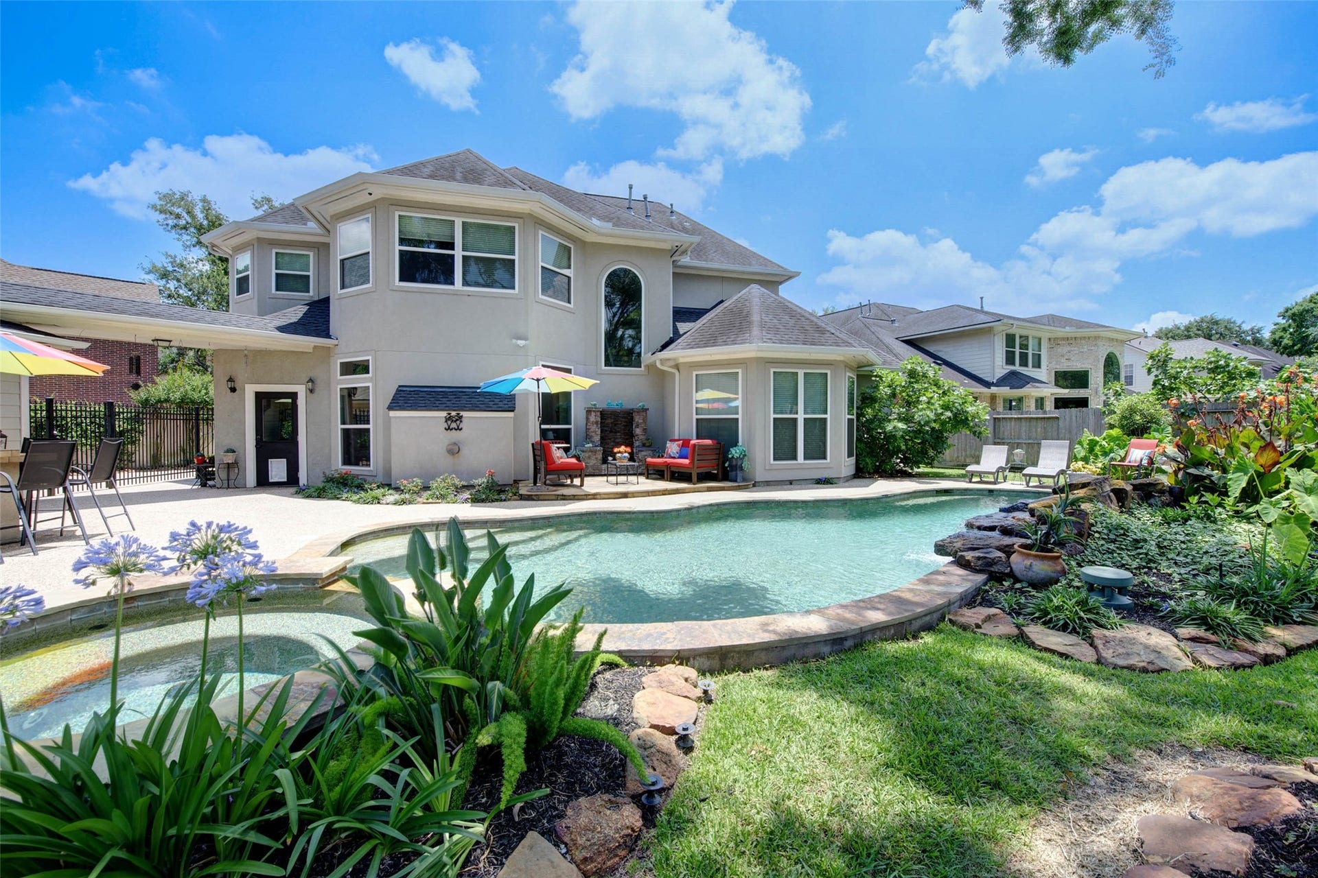 This photo showcases a spacious backyard featuring a beautiful swimming pool surrounded by lush landscaping. The two-story house has a modern design with large windows, offering plenty of natural light. There's a cozy outdoor seating area with umbrellas, perfect for relaxation and entertaining.