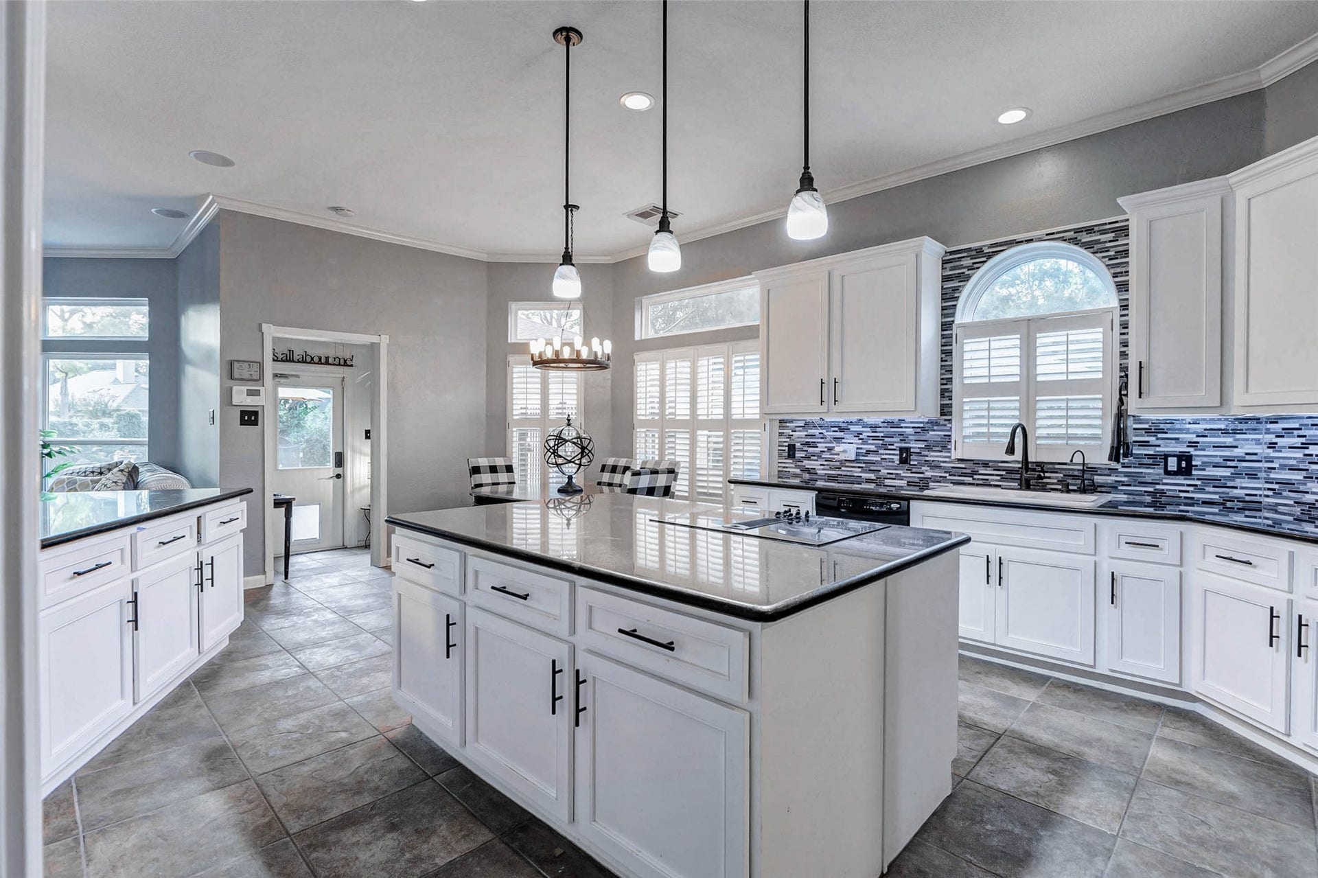 This modern kitchen features white cabinetry, a central island with granite countertops, and pendant lighting. It includes a stylish tile backsplash, stainless steel appliances, and ample natural light through arched windows with shutters. Views of the Mud room in the background.