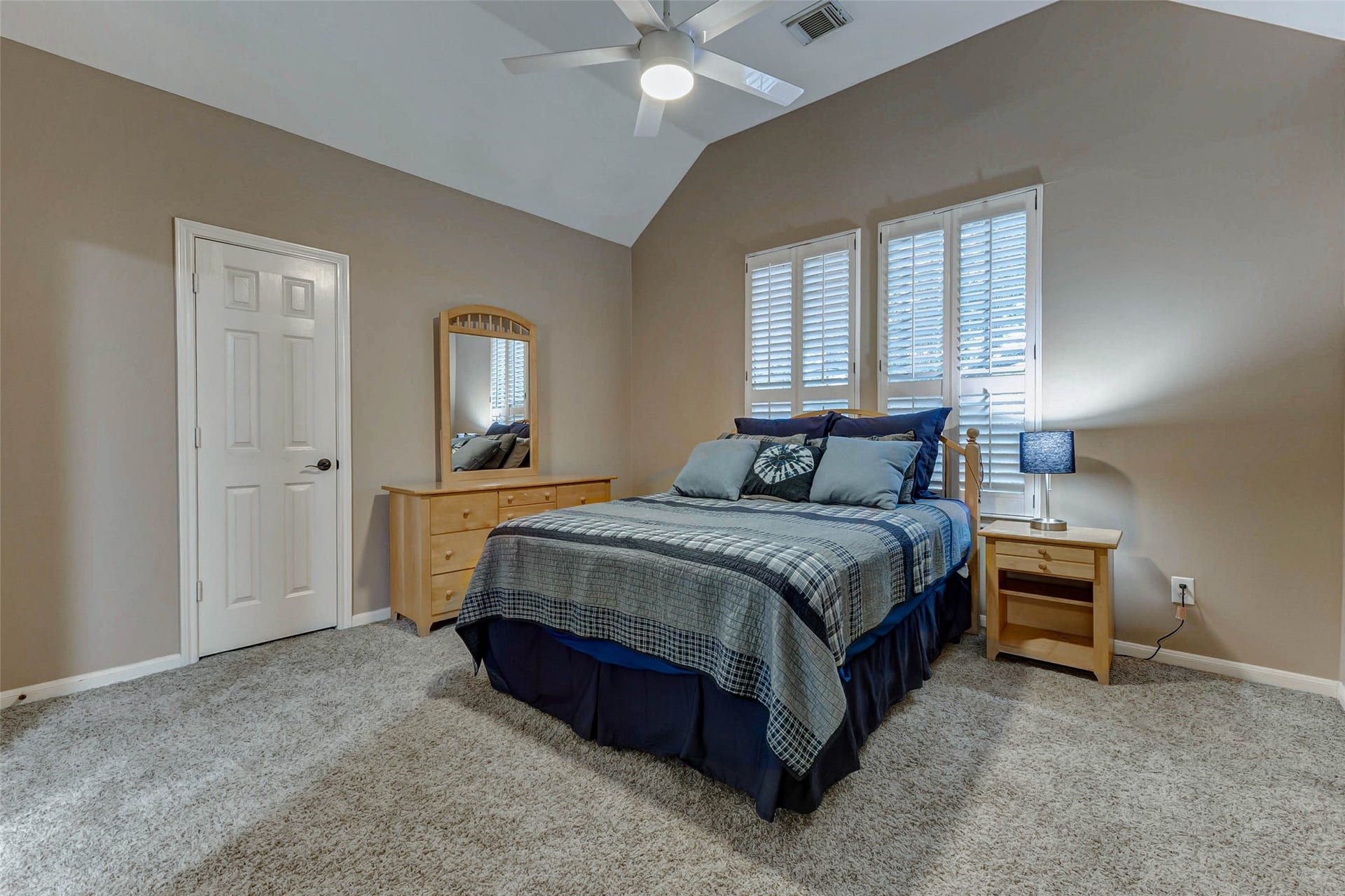 This cozy bedroom features a soft carpeted floor, neutral walls, and a high ceiling with a fan. Natural light filters in through the window with shutters, creating a warm and inviting atmosphere.