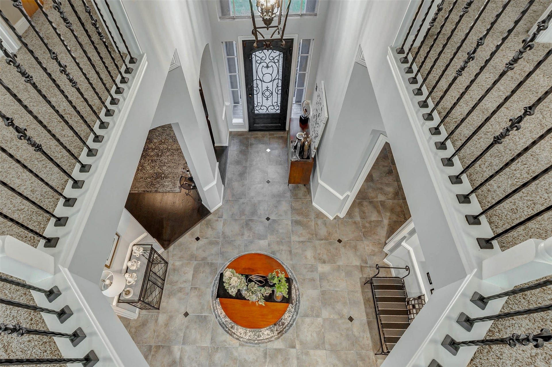This photo showcases an elegant foyer with 20' high ceilings and an overhead view of a grand entrance. The space features a decorative wrought iron staircase, tiled flooring, and a gorgeous chandelier. Space for a round table with floral arrangements adds a welcoming touch.