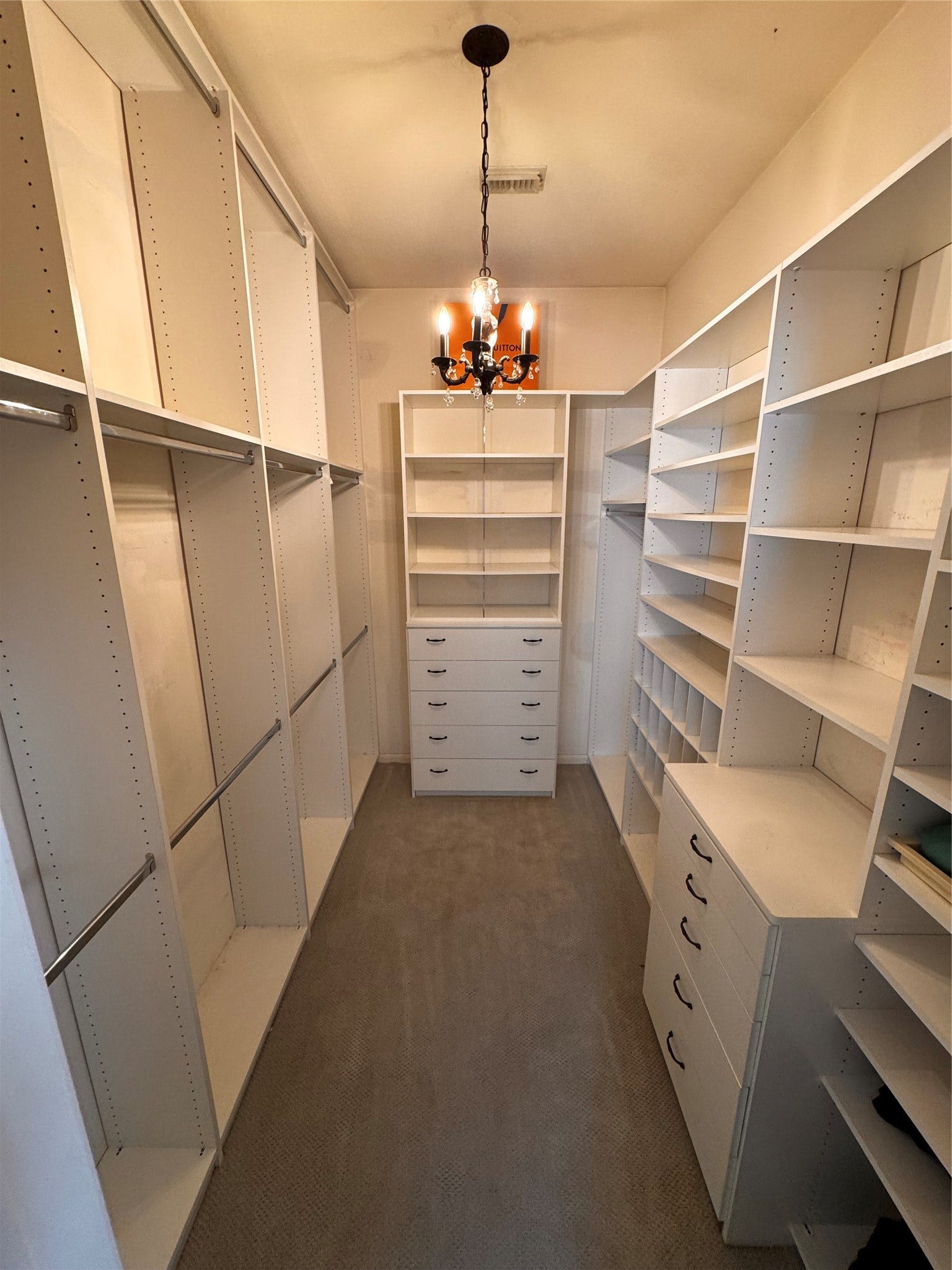 Spacious walk-in closet (there are 2) with custom shelving, hanging space, and drawers, featuring a chandelier for added elegance. Ideal for organized storage. Custom spacious walk-in closet, with shelving,  3 tiered hanging space, 2 sets of custom drawers, and space for 75 pairs of shoes, boots and sandals.
