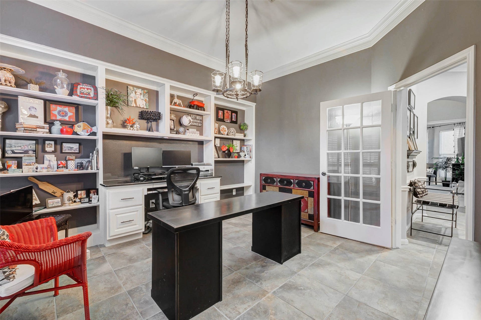 This home office features built-in shelving with ample storage and built-in desk with granite top.The room is well-lit with a decorative chandelier and has a modern, stylish design. French doors open to an adjacent room, creating a bright and inviting workspace.