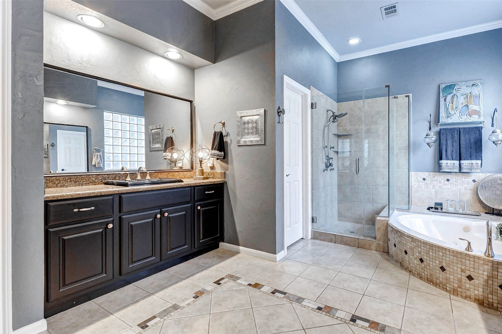 This bathroom features a spacious layout with a large vanity and double sinks, a glass-enclosed shower, and a corner soaking tub. The neutral tile flooring and soft wall colors create a serene atmosphere, complemented by ample lighting and elegant fixtures.