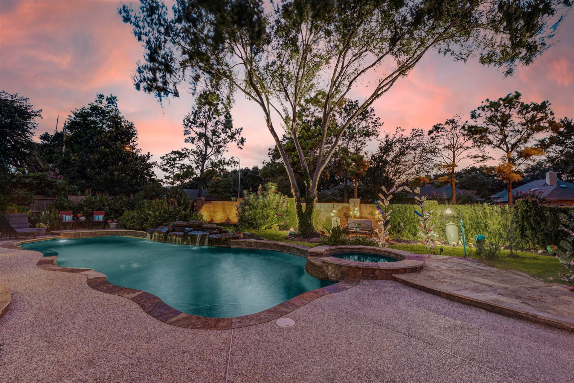 This backyard features a beautifully landscaped pool and spa, surrounded by lush greenery and mature trees. The stone patio offers a serene setting, perfect for relaxation or entertaining, with a backdrop of a colorful sunset sky.  8 outdoor plugs situated around the yard for outdoor lighting. Lighting stays with the house.