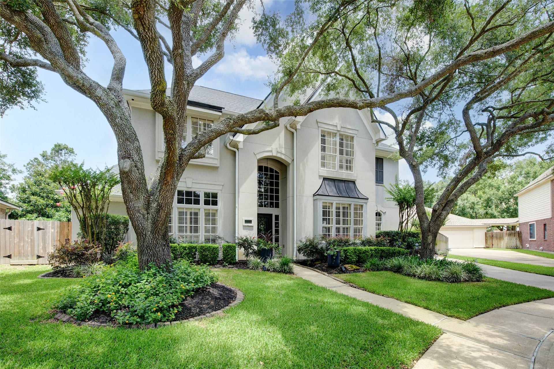 This elegant two-story home features a grand entrance with large windows and a beautifully landscaped front yard. Mature trees provide ample shade, and the driveway leads to a spacious garage. Ideal for those seeking a blend of style and comfort.