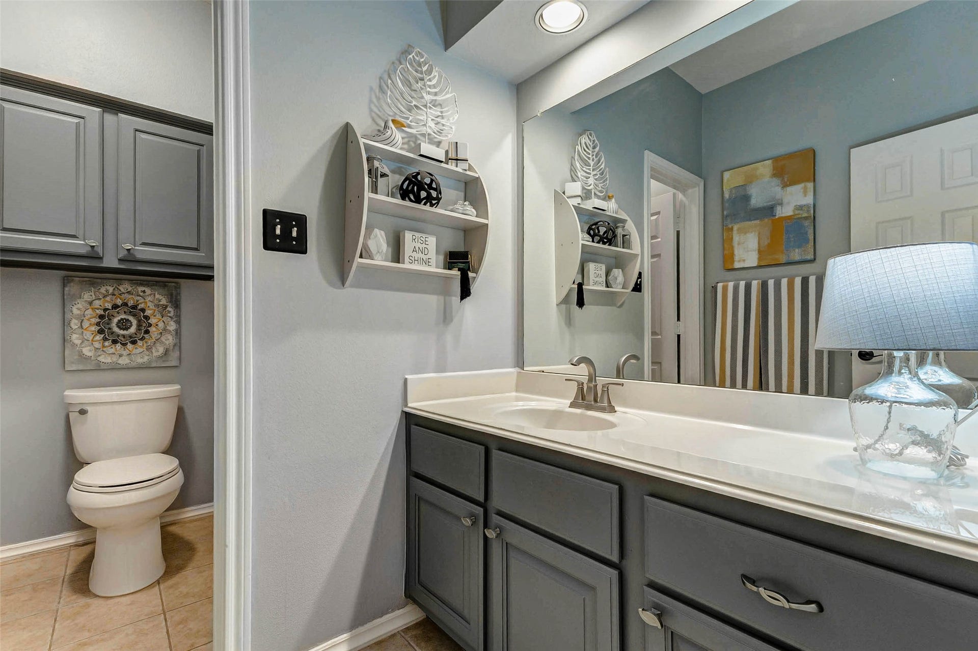 This bathroom features modern gray cabinetry with ample storage, a spacious vanity with a sleek countertop, and stylish decor. The room is well-lit with overhead lighting, it includes a separate toilet area with tile flooring for easy maintenance.