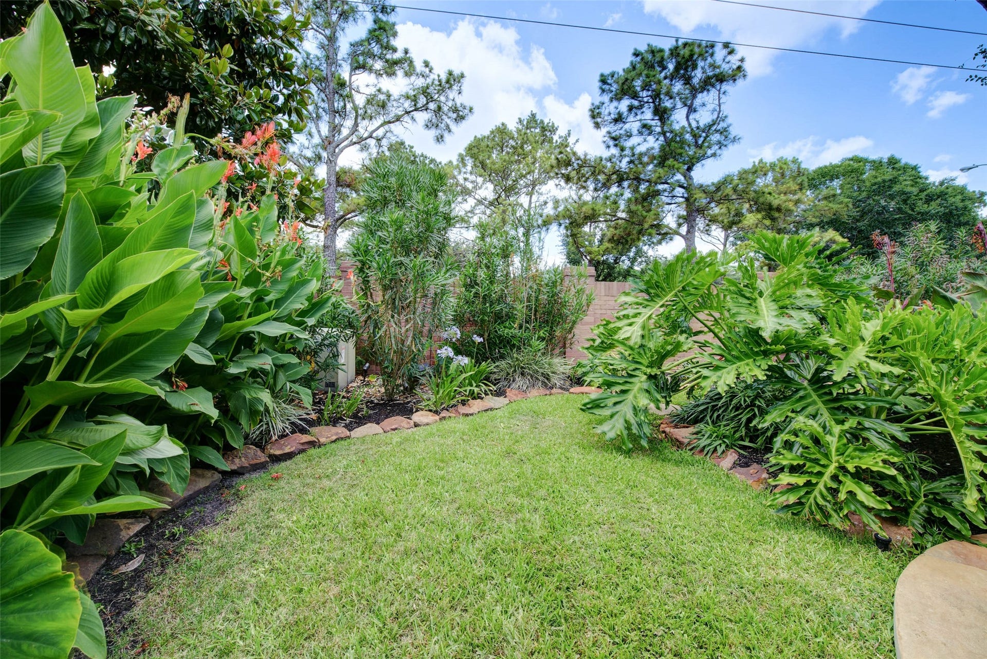 This photo showcases a lush, well-maintained backyard with vibrant greenery and tropical plants, bordered by stone edging. The space offers a serene outdoor setting, perfect for relaxation. Room for kids play house, trampoline or other play structures. Grass goes all the way around the pool.