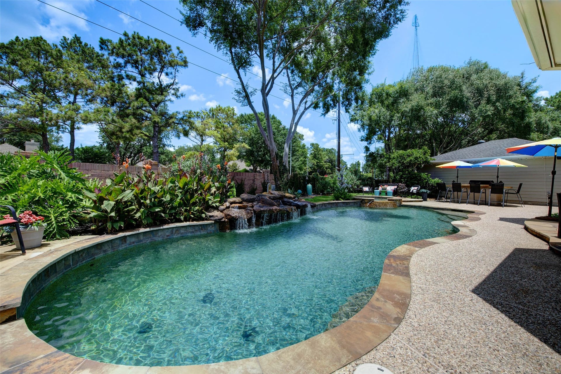 This photo showcases a beautiful backyard with a large, curved swimming pool featuring a waterfall. The pool is surrounded by lush greenery and a spacious patio area with seating and colorful umbrellas, providing a perfect setting for relaxation and entertainment.