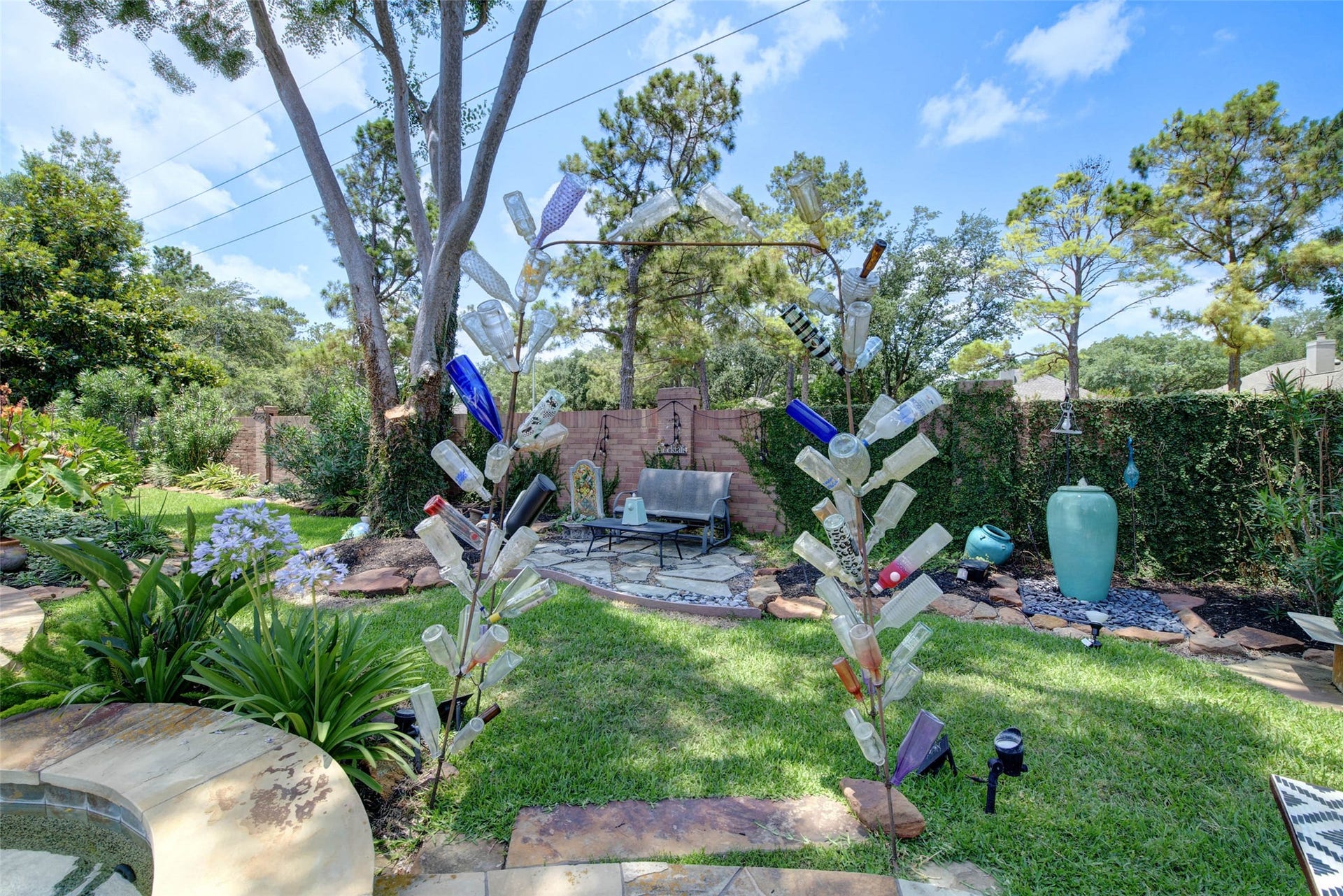 This backyard features a vibrant garden with lush greenery and mature trees. It includes a cozy seating area on a stone patio, and decorative elements like a large ceramic vase fountain. The space is perfect for relaxation and outdoor entertaining. The two fountains stay with the house.