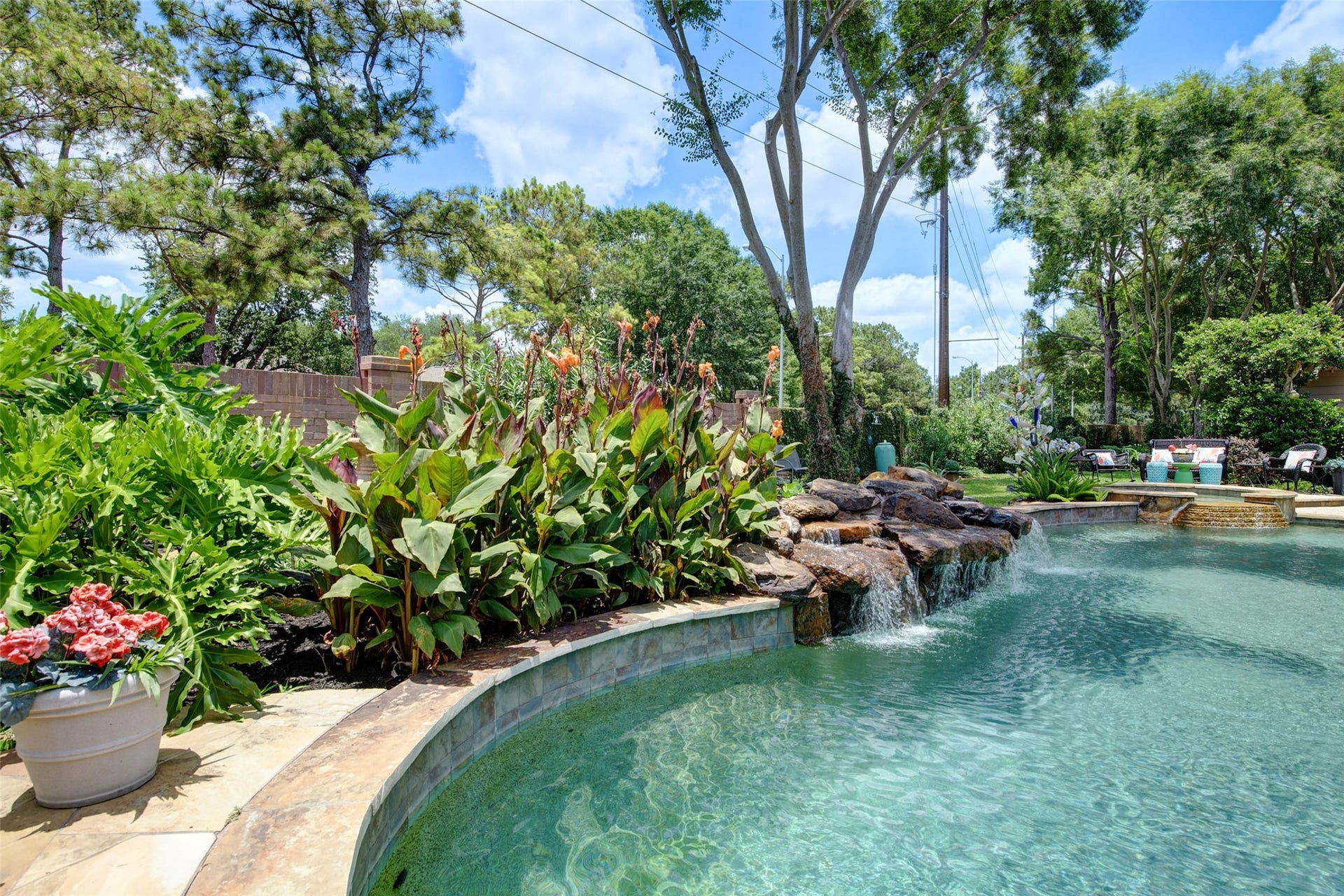 This photo showcases a beautifully landscaped backyard with a clear blue swimming pool featuring a rock waterfall. Surrounded by lush greenery and vibrant flowers, the setting offers a serene and inviting outdoor space perfect for relaxation and entertaining.