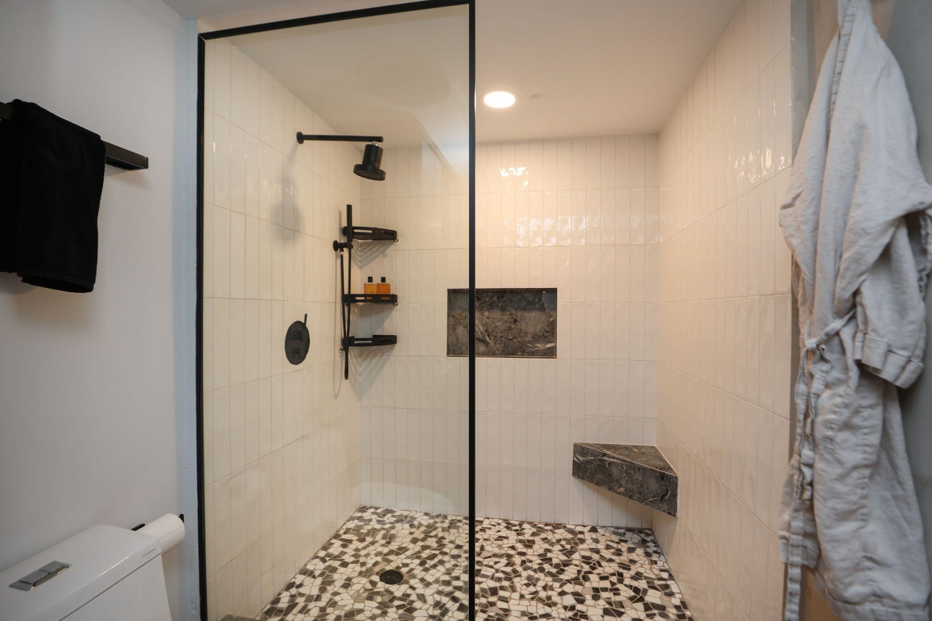 Oversized walk-in shower with a rainfall head, vertical subway tile, pebble-mosaic floor, matte black fixtures, built-in stone bench and a recessed niche for storage.