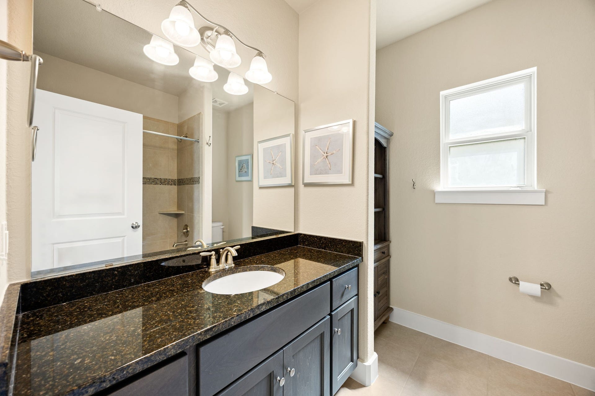 14 Kendrick Pines Boulevard, The Woodlands, TX 77389 This modern guest bath combines sleek design with everyday functionality. A deep navy vanity is paired with a polished granite countertop, creating a sophisticated focal point against the neutral walls. A large mirror and bright vanity lighting enhance the space, while the tiled shower and natural light from the privacy window ensure a bright, clean, and inviting atmosphere for guests.