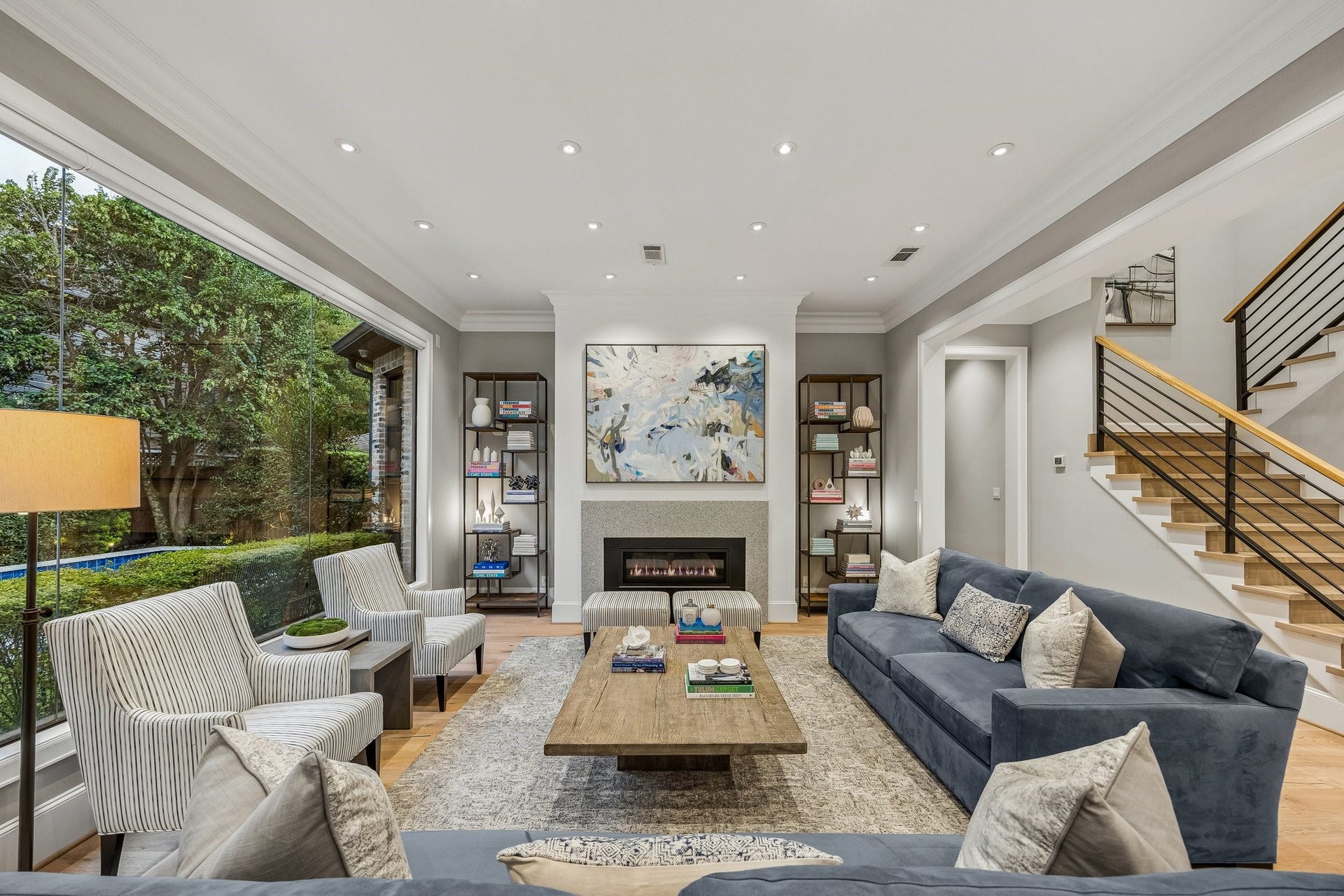 This beautifully appointed living room seamlessly blends contemporary sophistication with timeless architectural elegance. A dramatic floor-to-ceiling picture window serves as the rooms focal point, offering breathtaking views of the lush, meticulously landscaped yard while filling the space with natural light and creating a seamless connection between indoor and outdoor living.