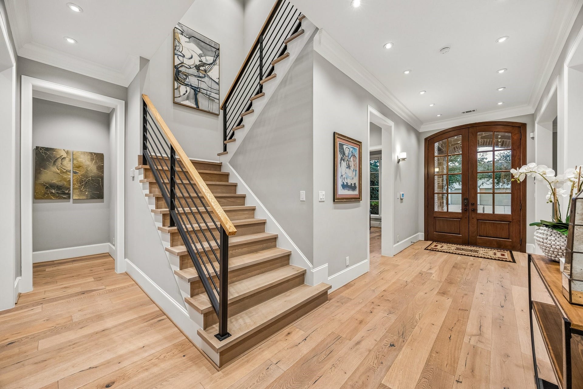 This stunning entry foyer showcases a beautiful blend of contemporary architectural design and timeless elegance. A striking staircase serves as the architectural focal point, featuring natural wood treads, sleek black horizontal iron railings, and a coordinating wood handrail, creating a sophisticated contrast of materials and textures.