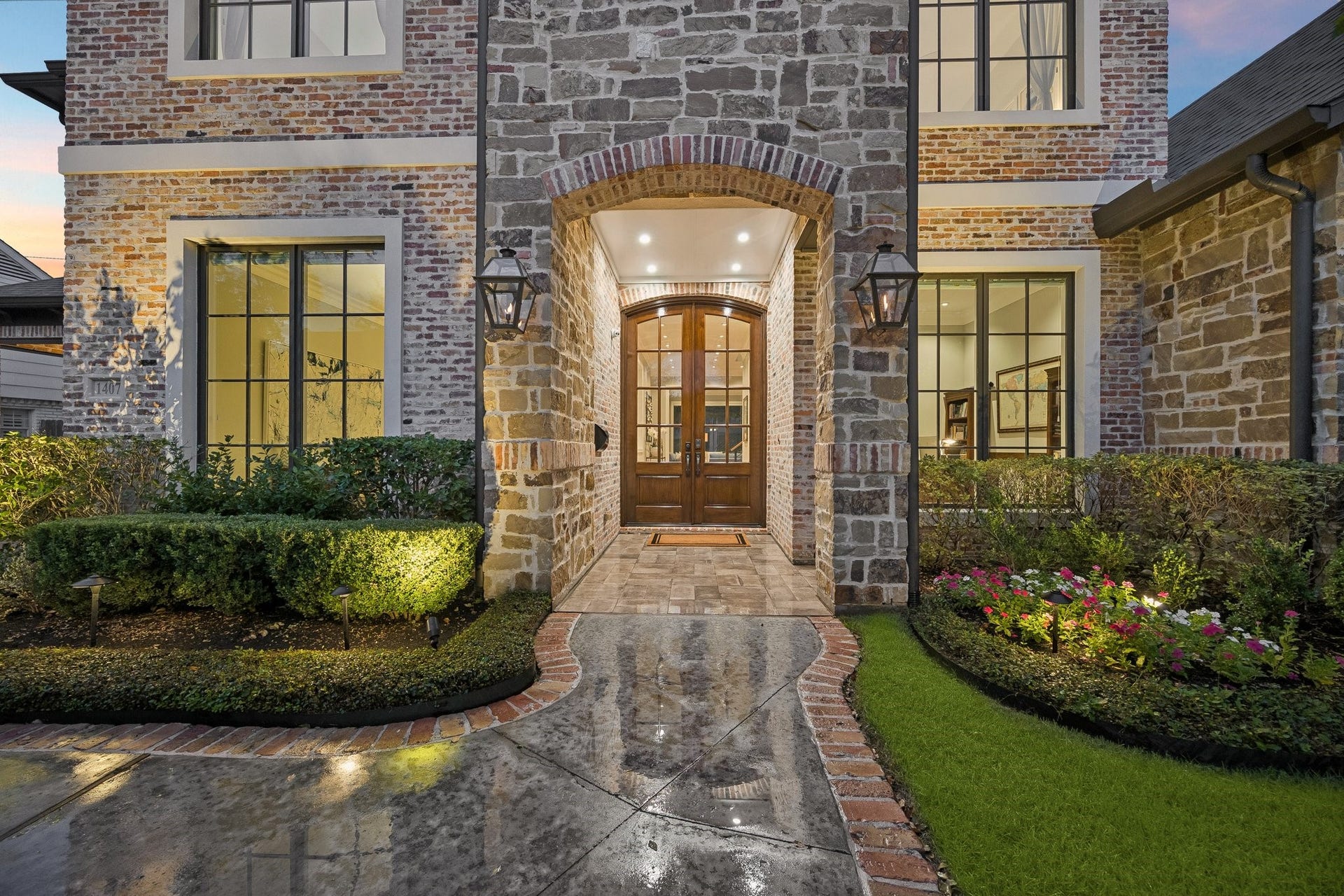 A charming, curved walkway bordered by brick leads through lush, manicured grounds to an impressive entryway featuring double wood-and-glass doors framed by a distinctive brick arch. Expansive windows invite natural light into the home, while warm exterior lighting beautifully accentuates the architectural features and creates an enchanting ambiance at dusk.