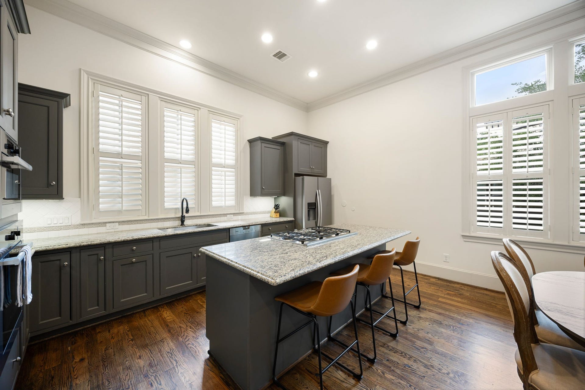 310 W Bell Street, Houston, TX 77019 This kitchen features elegant custom painted cabinetry, a spacious island with a built-in cooktop, and modern stainless steel appliances. Bright and airy, it has large windows with shutters and warm hardwood floors, creating a welcoming atmosphere.