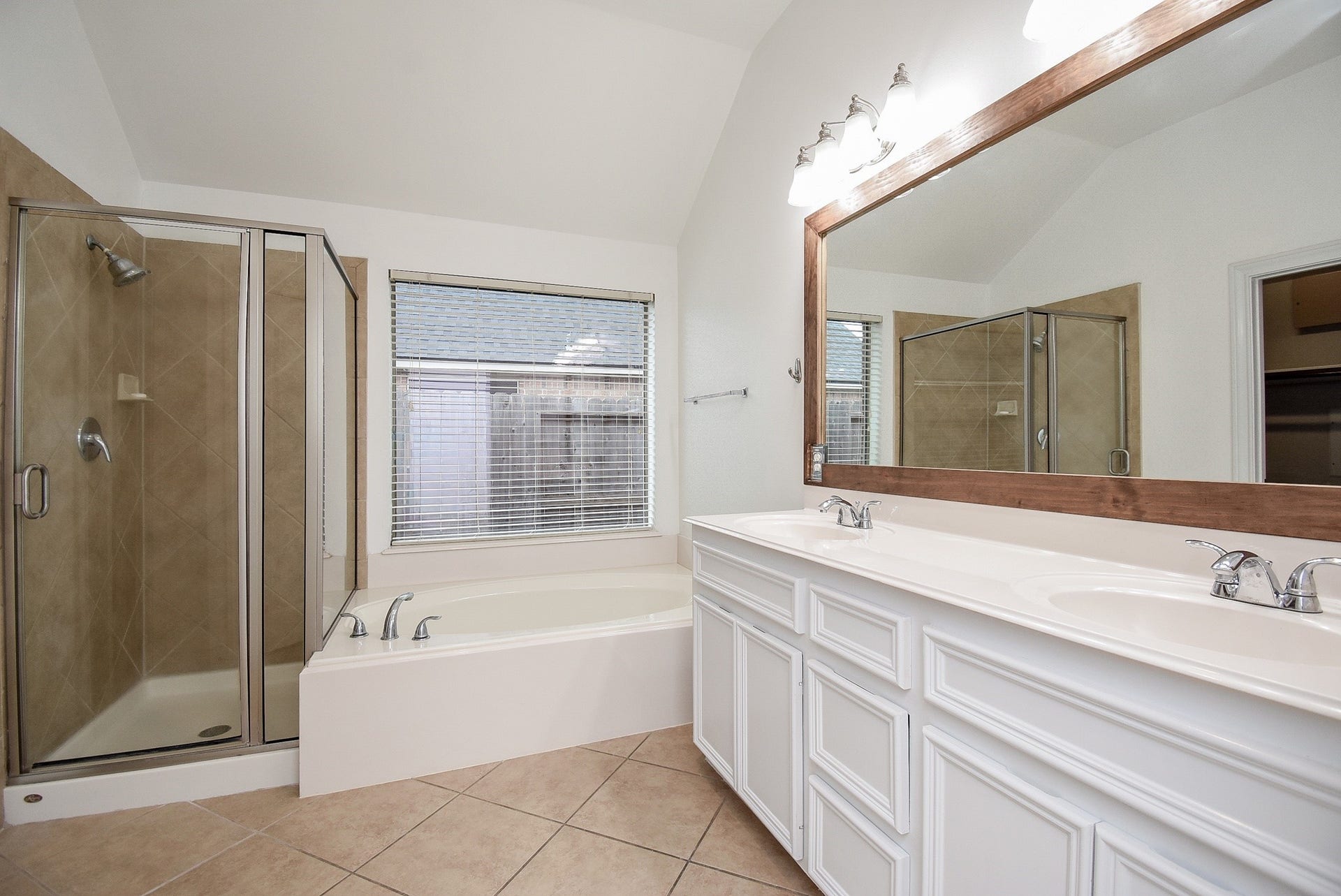 Unwind in the spacious primary bathroom featuring a dual-sink vanity with abundant counter space, a relaxing soaking tub perfectly positioned beneath a large window, and a separate glass-enclosed walk-in shower. Neutral tile flooring, generous natural light, and an oversized framed mirror create a bright, spa-like atmosphere, while the thoughtfully designed layout offers both comfort and functionality for everyday living.