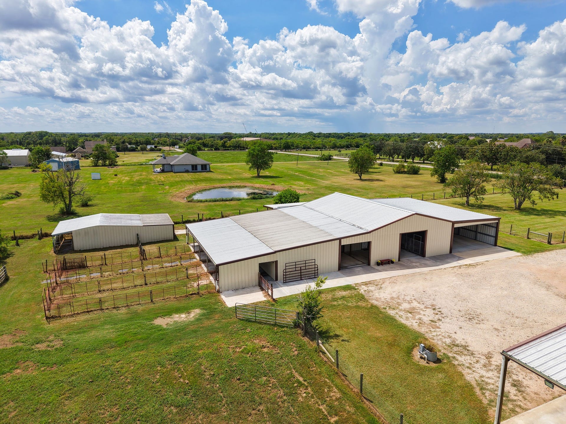 Expansive rural property featuring a spacious metal barn and multiple fenced paddocks, set amidst lush green fields and a serene pond, perfect for equestrian or agricultural pursuits.