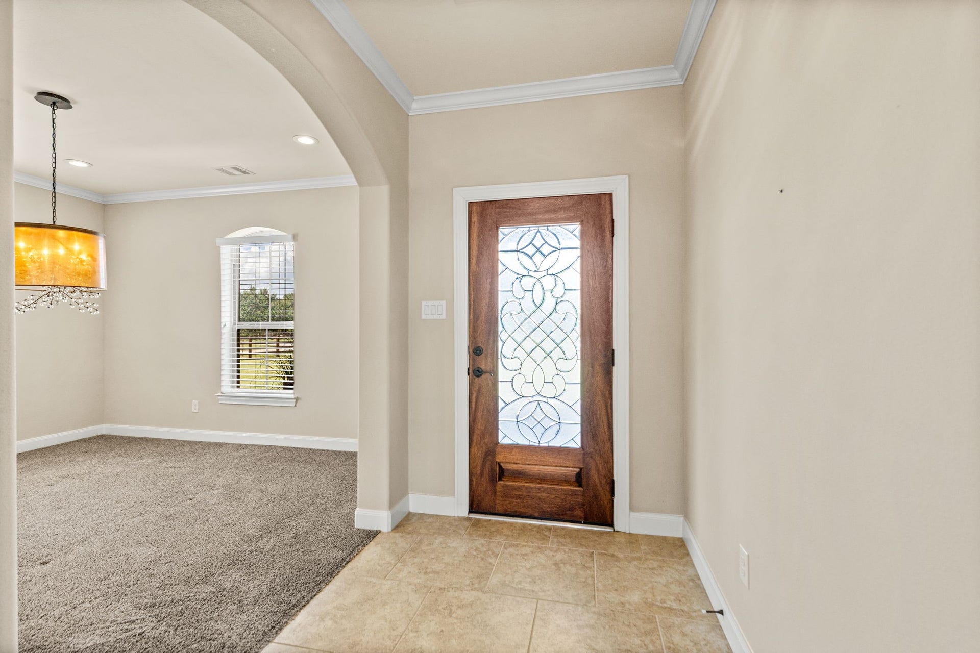 Elegant entryway featuring a beautifully crafted wooden door with intricate glass detailing, leading into a spacious carpeted living area with stylish lighting.